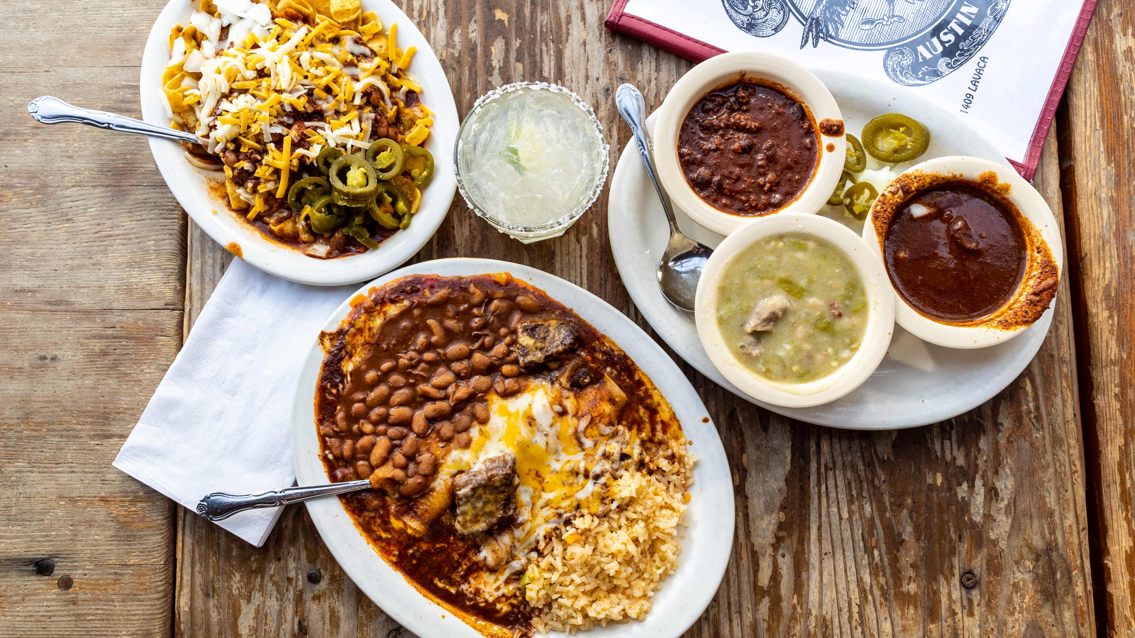 The Freidas Enchiladas, Frito Pie, Chili Sampler, and a Mad Dog Margarita sitting on a wooden table. A menu is set to the side.