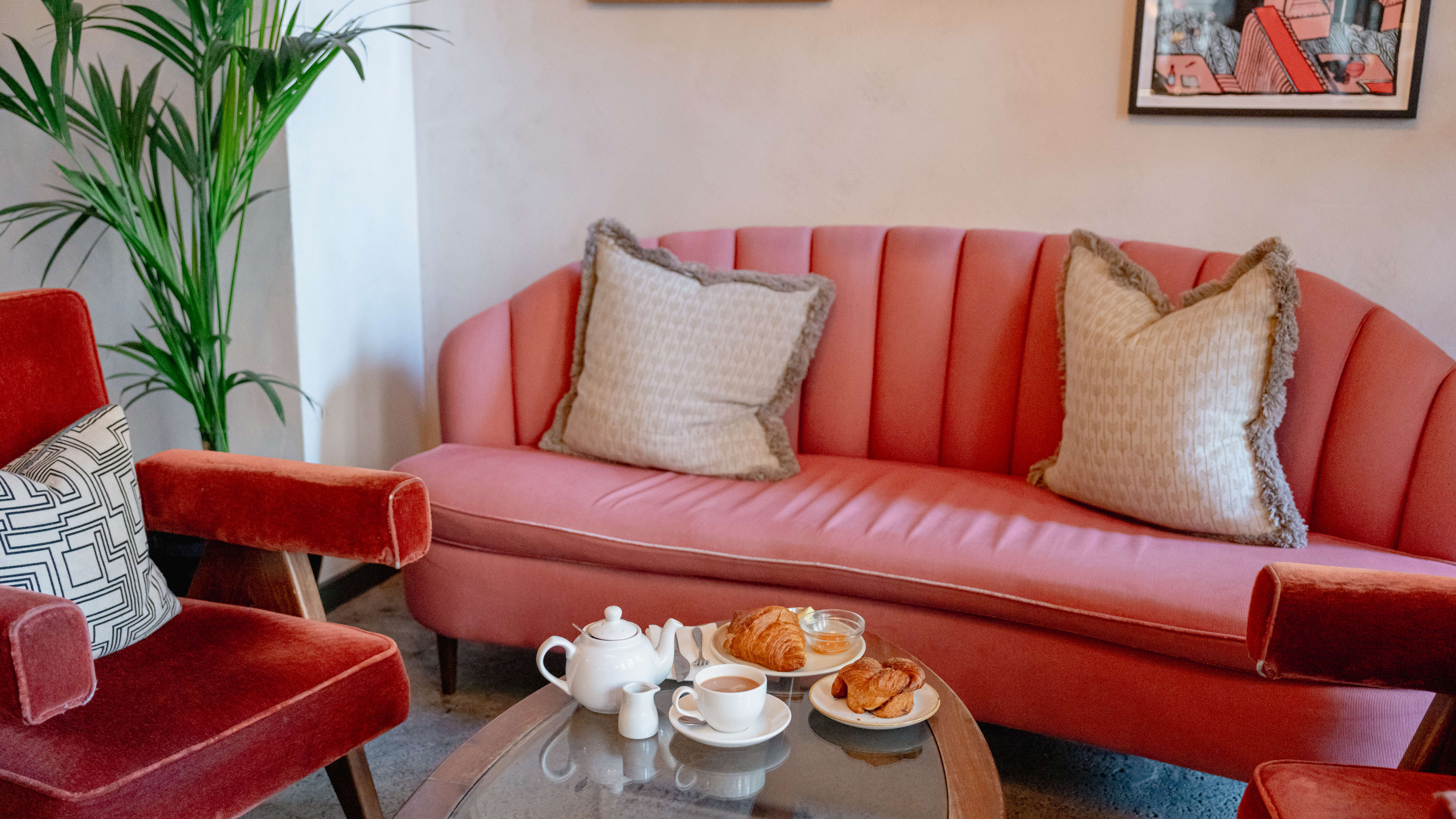 A pink velvet couch with beige patterned pillows. Two pink velvet chairs flank a glass coffee table with a teapot, cup of tea, and a few pastries.
