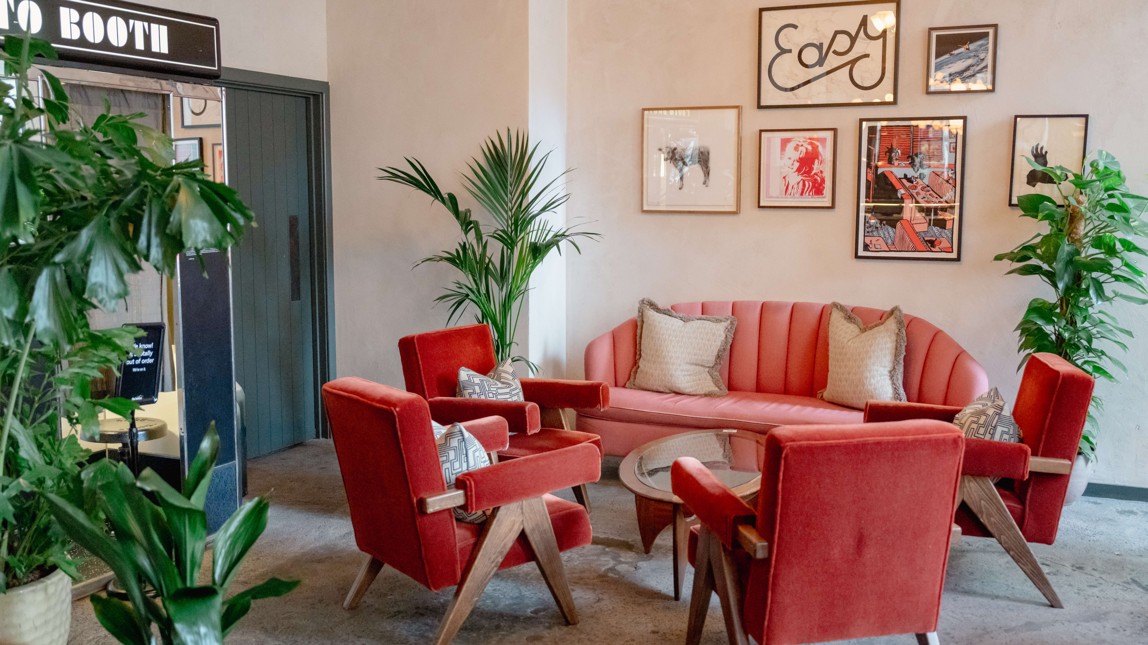 The interior of The Hoxton with a pink couch and four pink velvet chairs. There are various green plants and framed photos on the wall