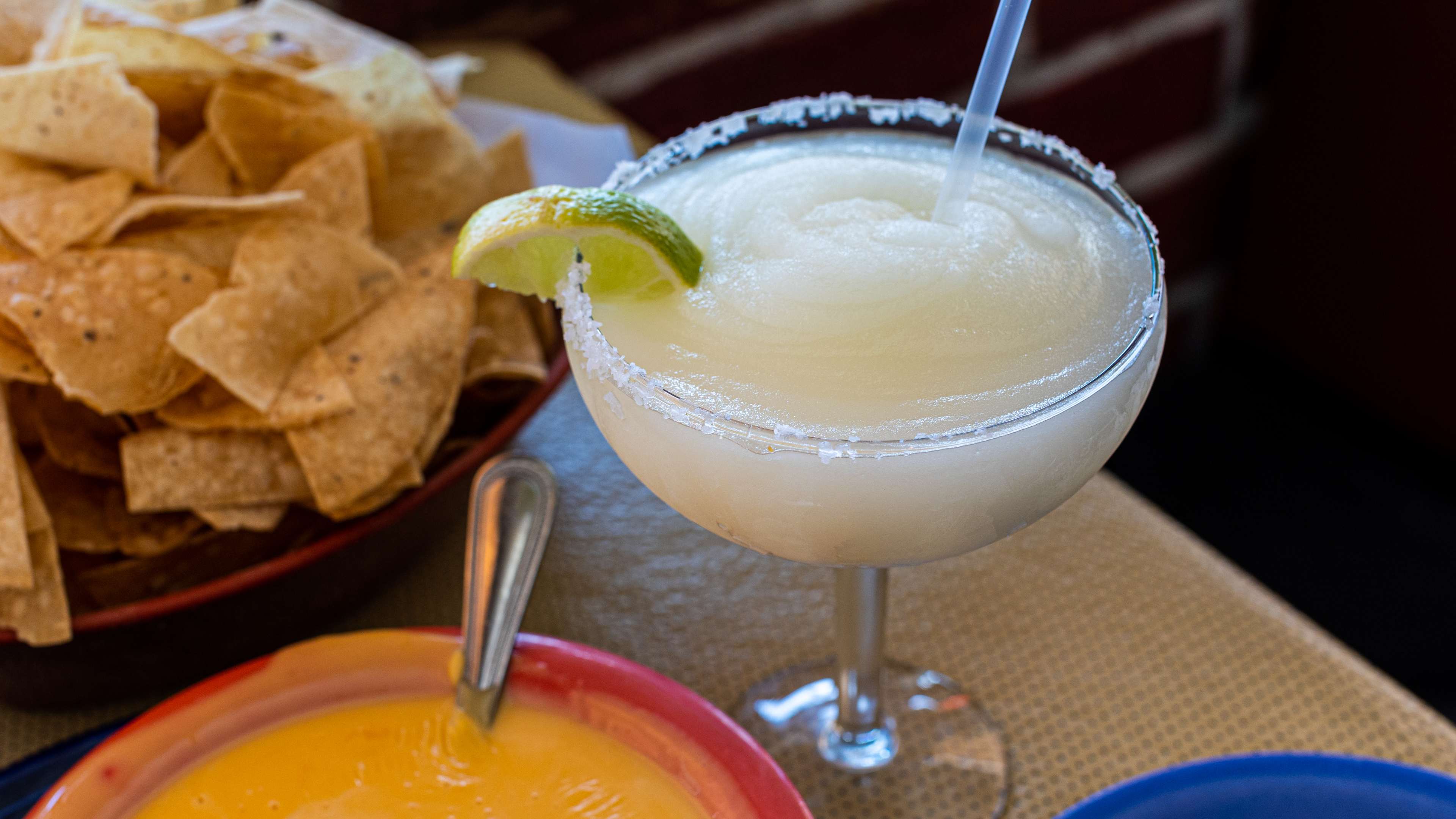 close up og a glass goblet full of a frozen margarita cocktail with a salted rim, wedge of lime, and clear straw on a table with a basket of chips and bowl of queso