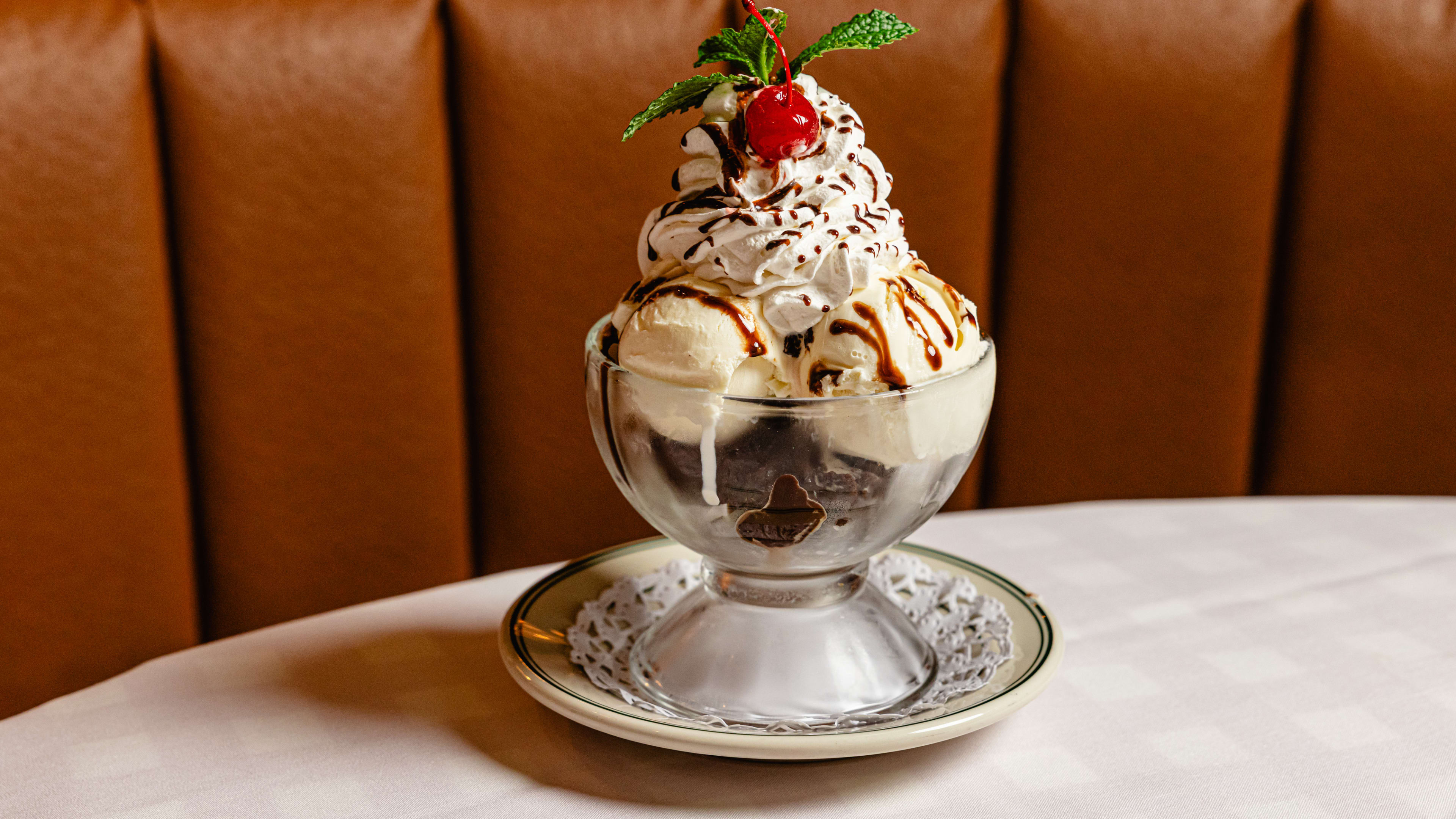 An ice cream sundae with whipped cream and a cherry, served in a glass dish on a table covered in a white tablecloth.