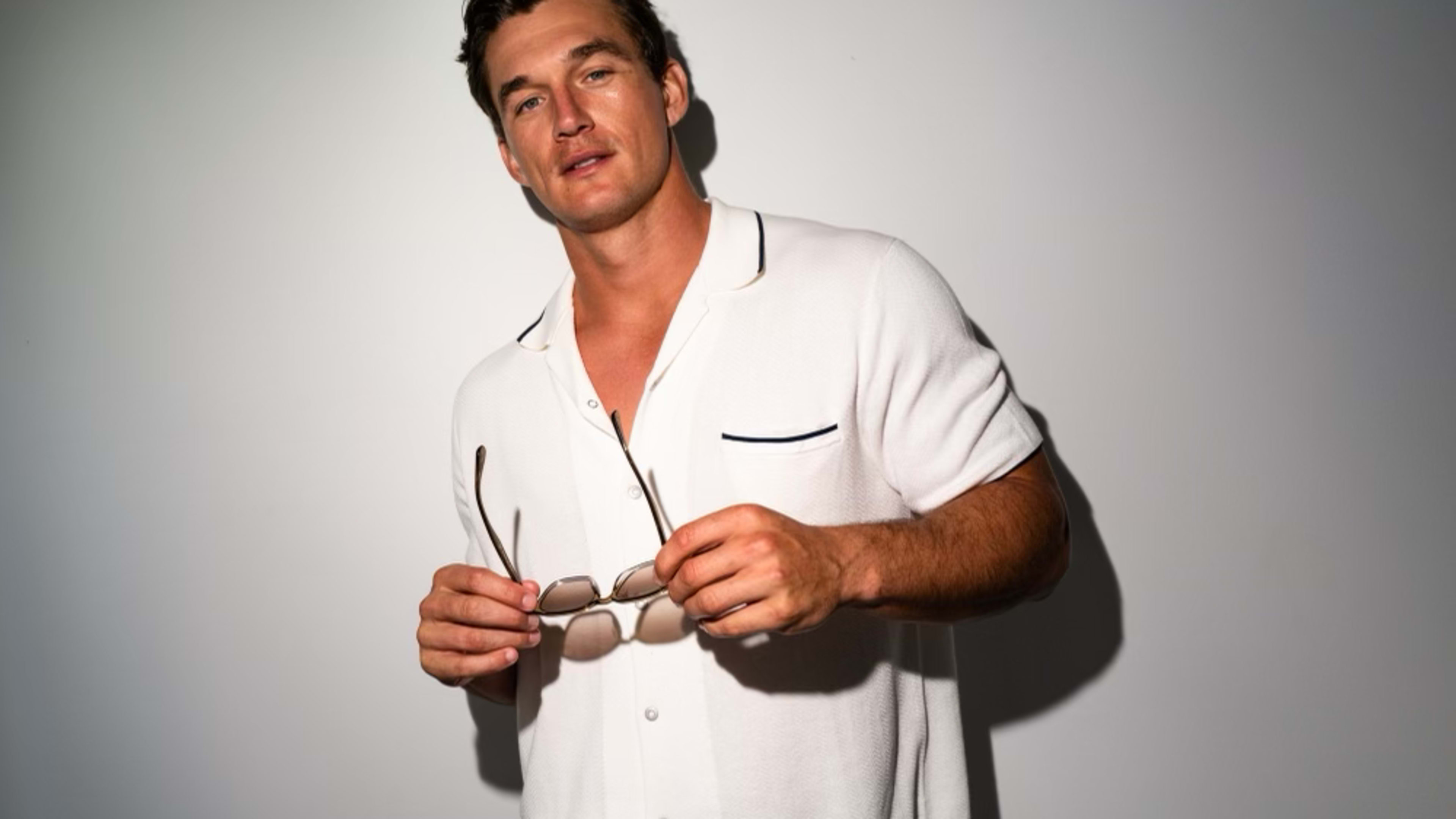 former Bachelorette contestant Tyler Cameron wears a white polo shirt and blue jeans and holds a pair of sunglasses in his hands