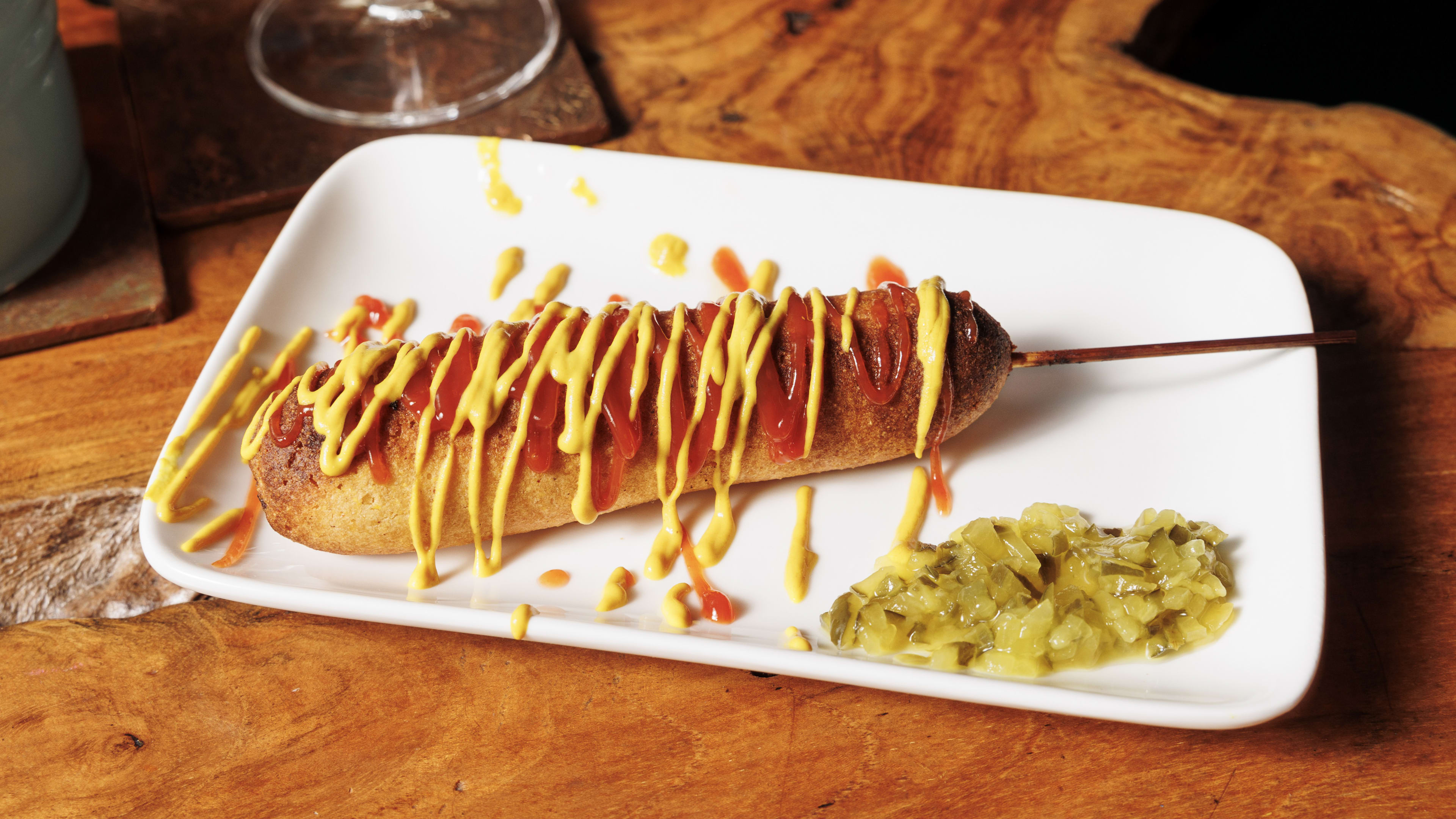 Corn dog drizzled with ketchup and mustard on a plate next to relish.