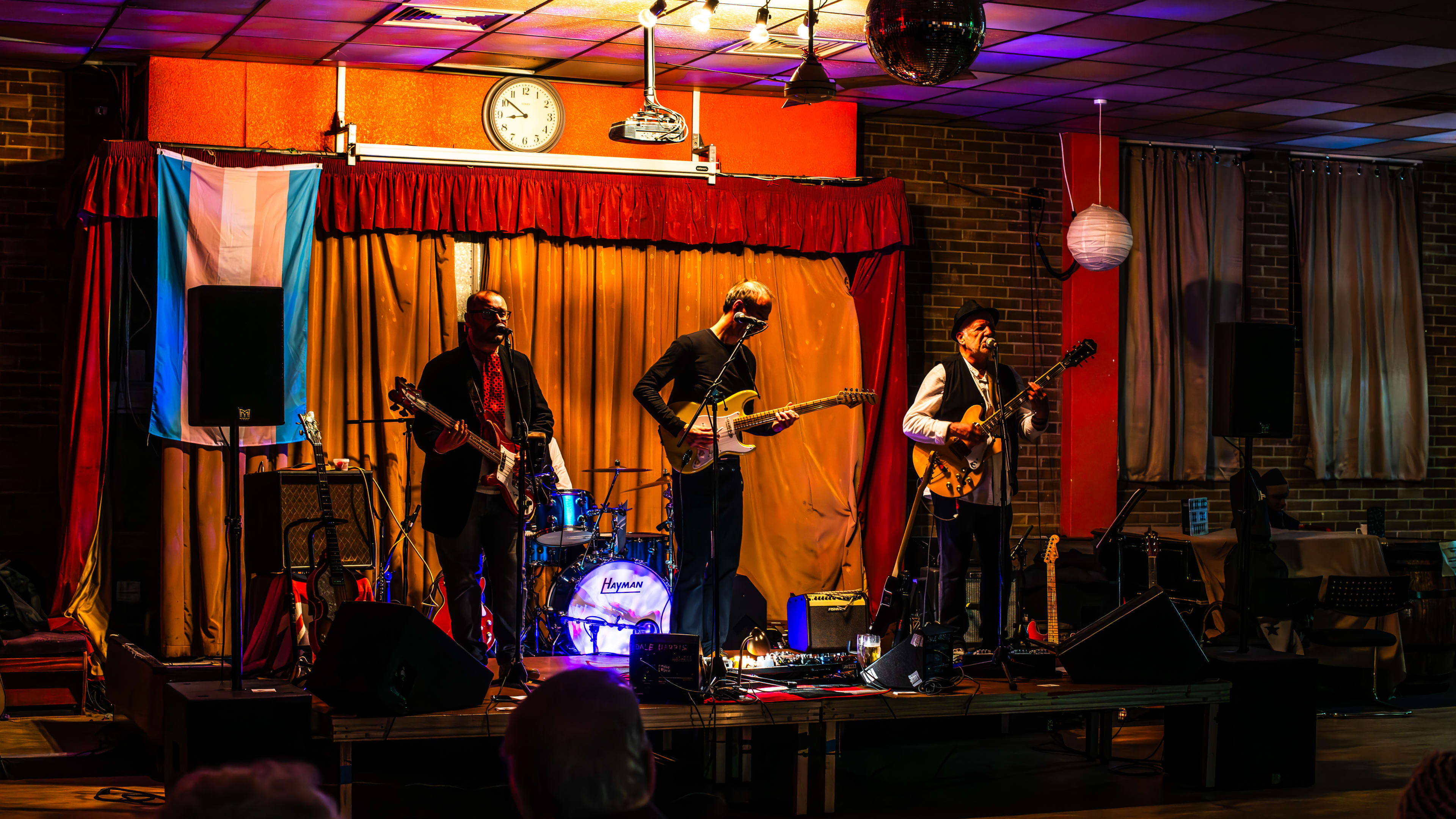 Three people performing on stage at Walthamstow Trades Hall. They are all holding guitars and singing at mics.  There is a trans flag hanging behind them as well as an orange and red curtain.