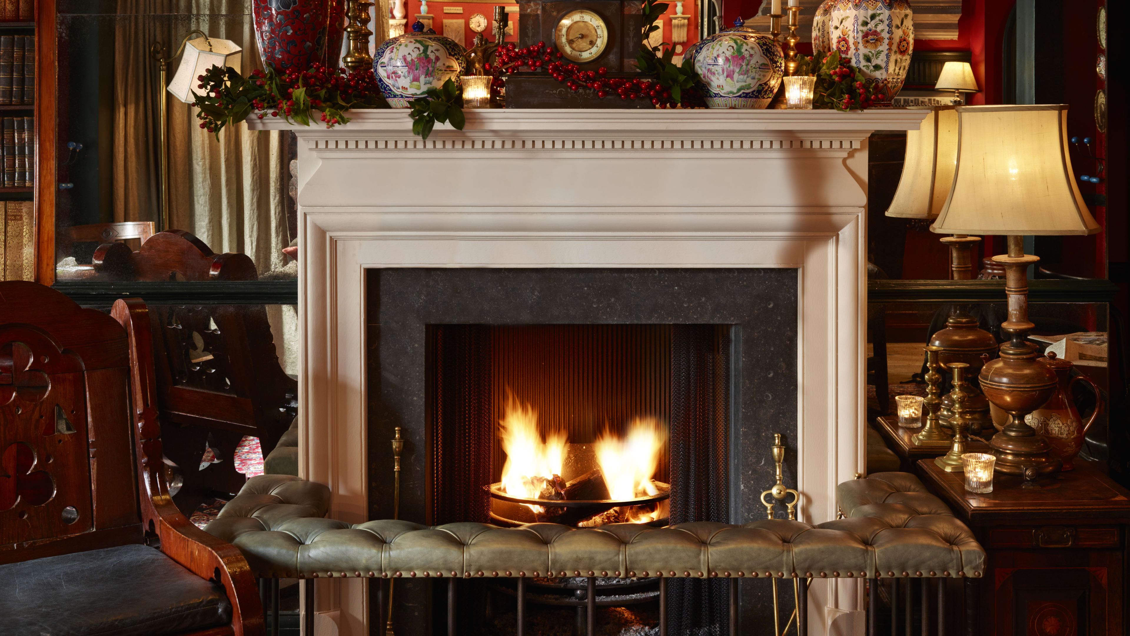 A lit fireplace with a wrap-around bench, side table with a lamp, wooden chair, and a mantel with trinkets, a clock and some plants.