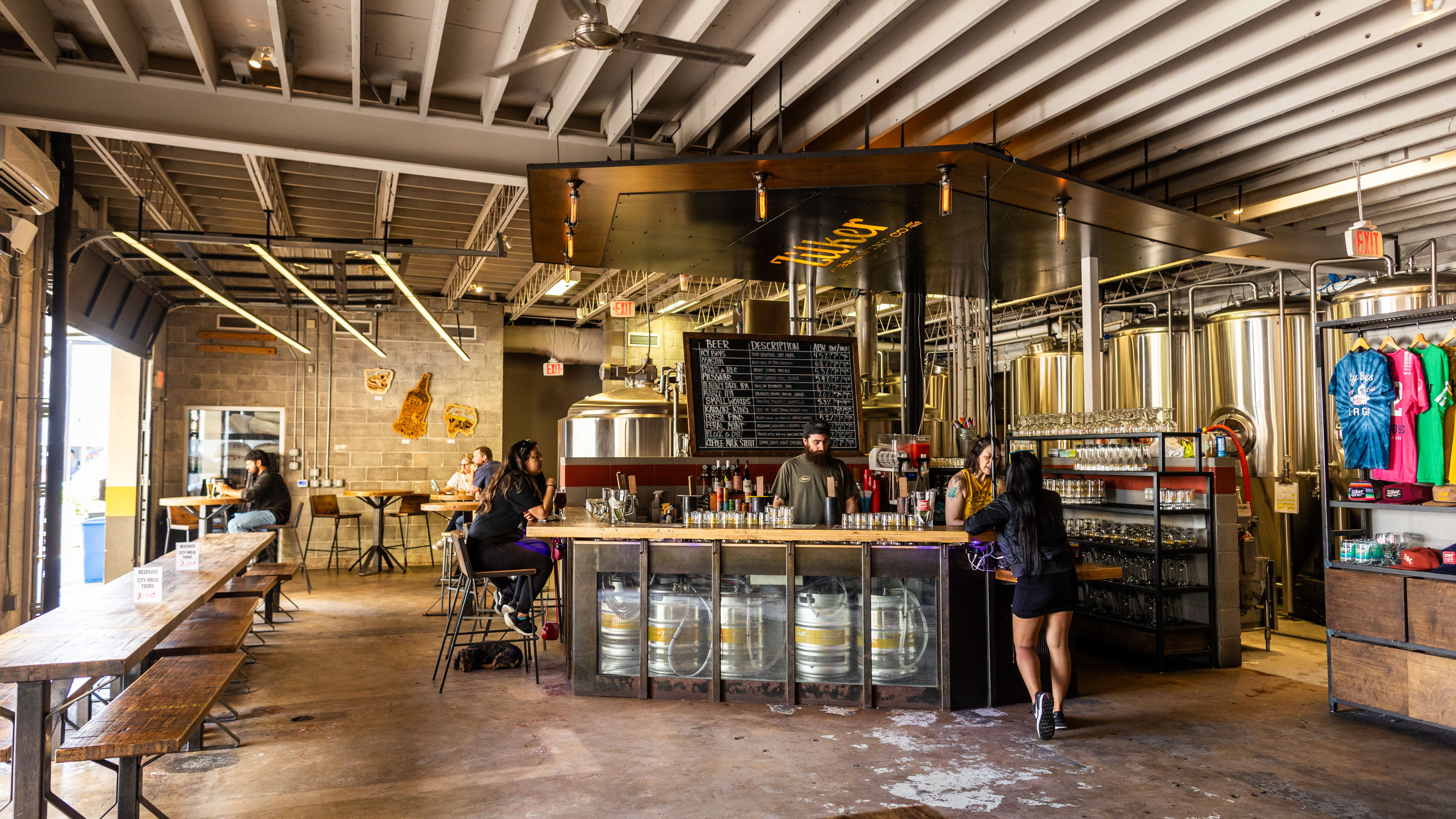 The interior of Zilker Brewing with open garage doors to the patio, metal brew barrels and kegs, wooden counter seating next to the windows and merch on shelves.