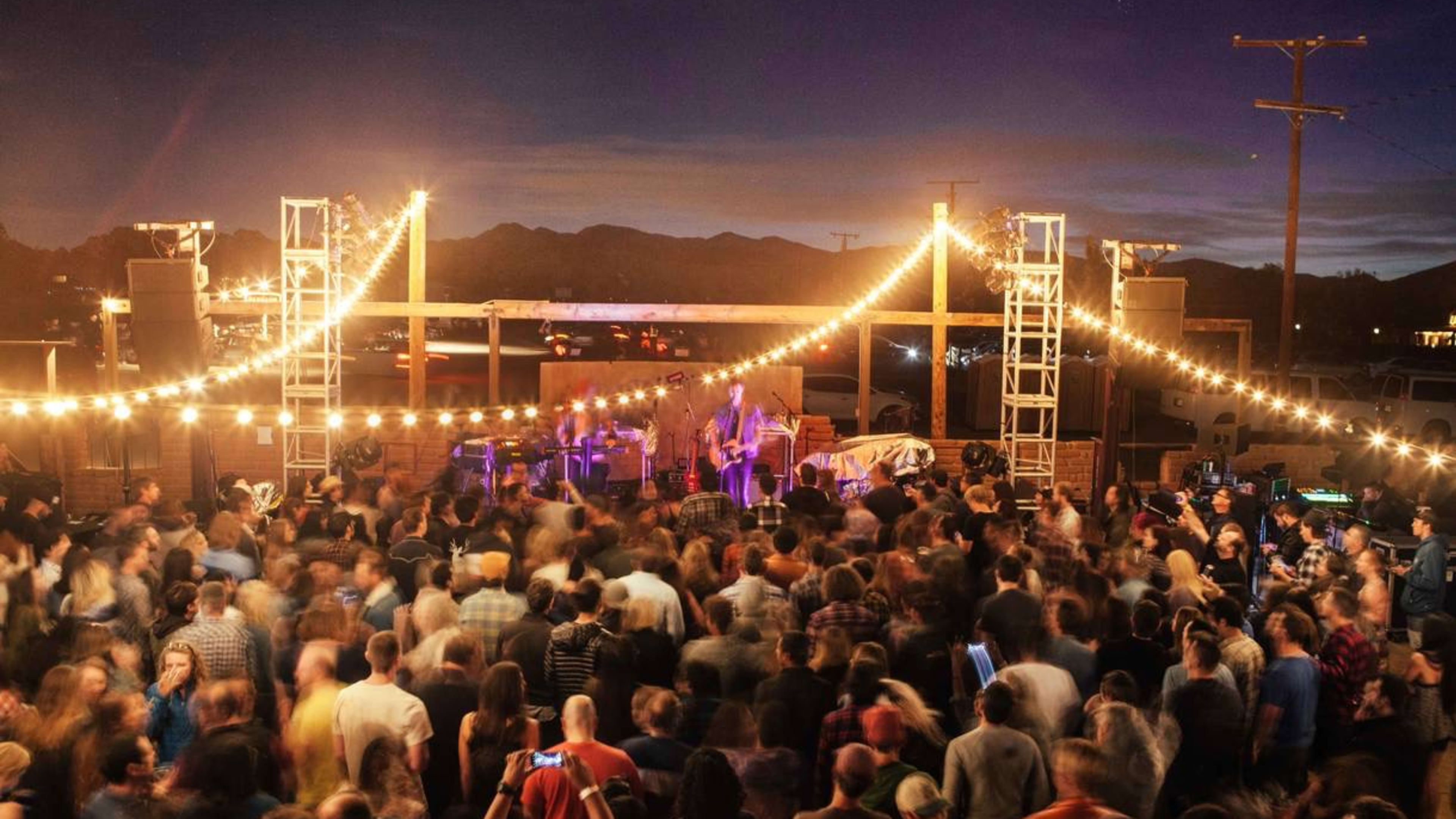 musicians on a small outdoor stage performing for a large crowd under hanging twinkle lights.