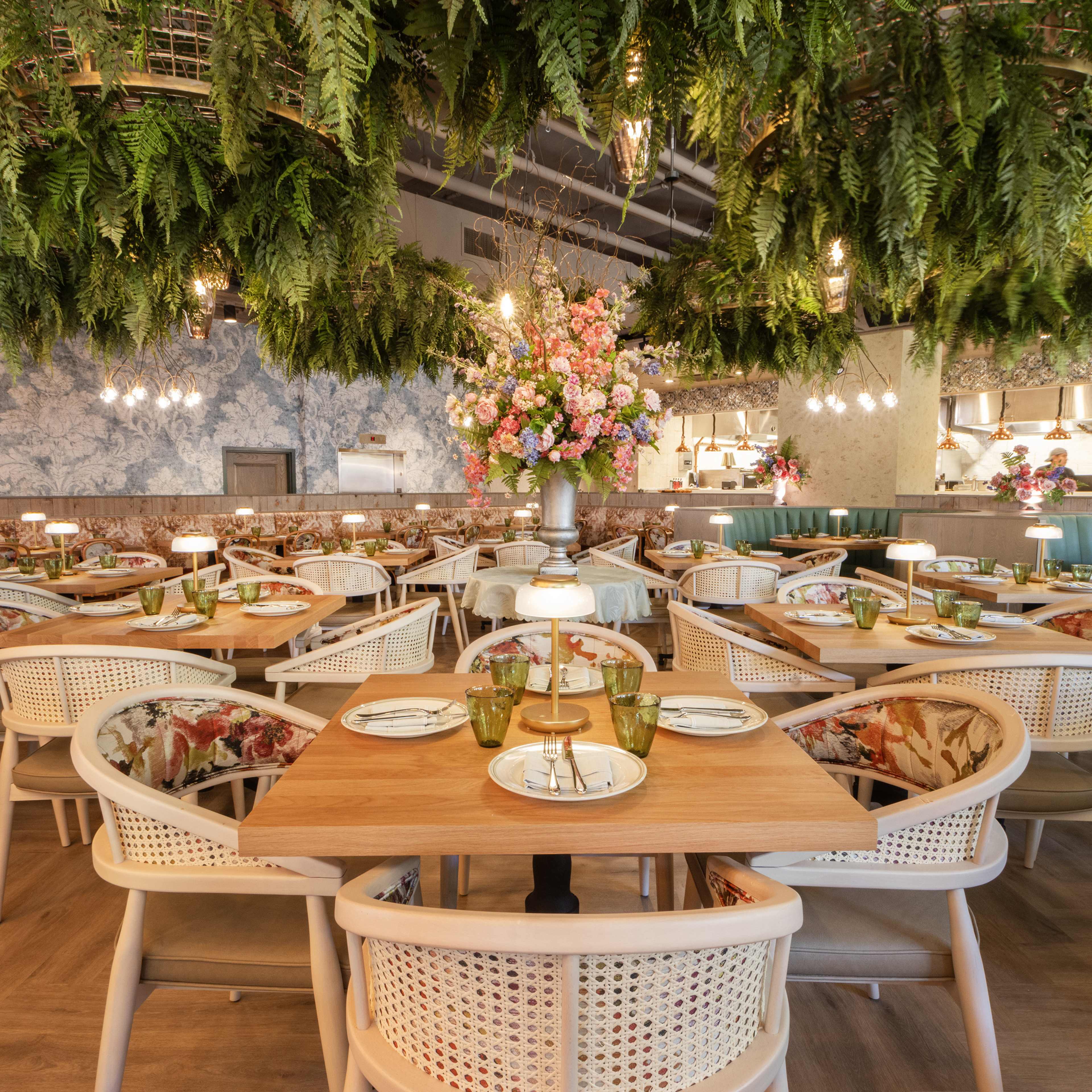 A bright dining room full of tables with plants hanging from the ceiling and an open kitchen in the background.