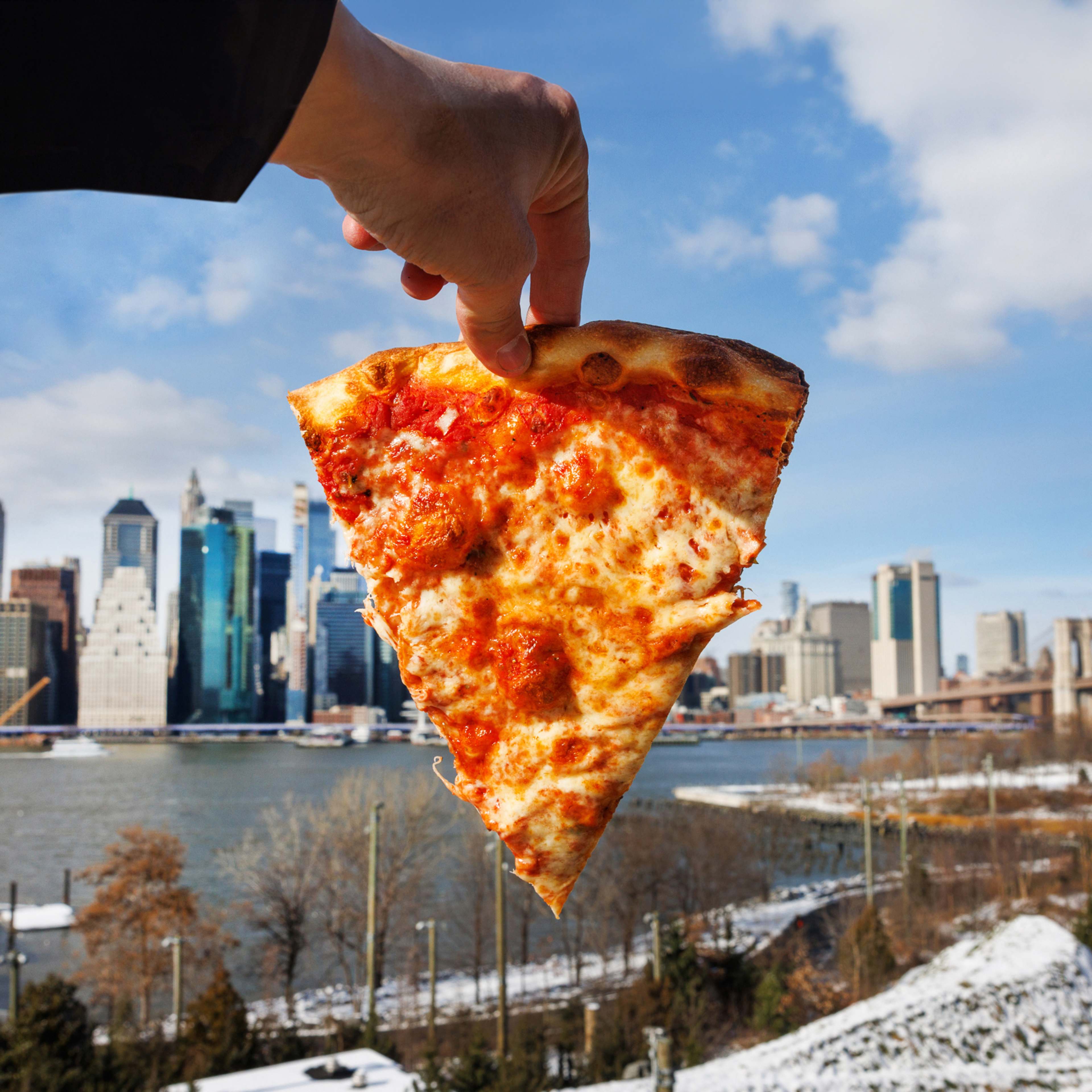 A slice of cheese pizza held up against the NYC skyline. There is a light dusting of snow on the ground.