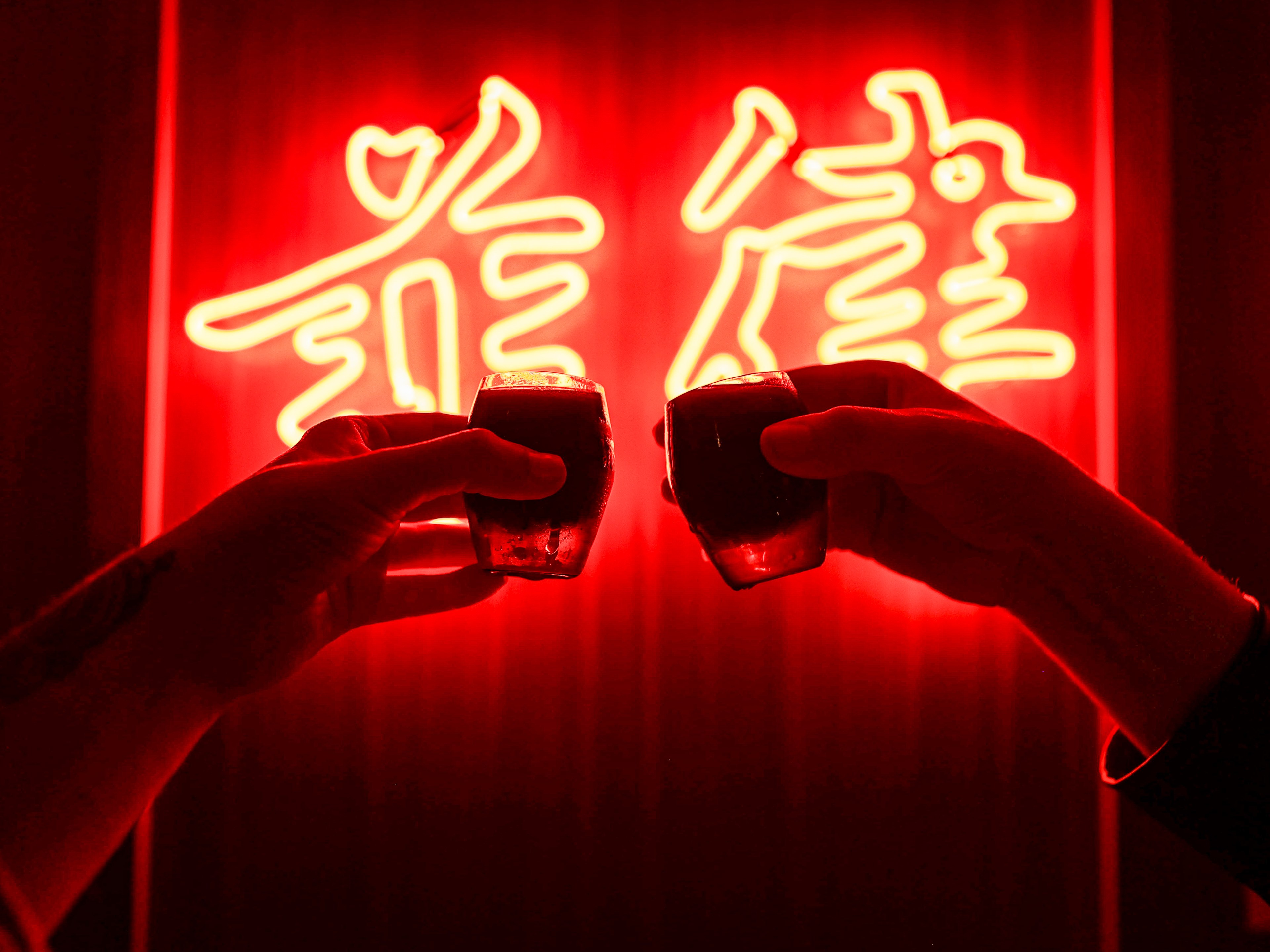 two people holding up cocktails in front of neon sign