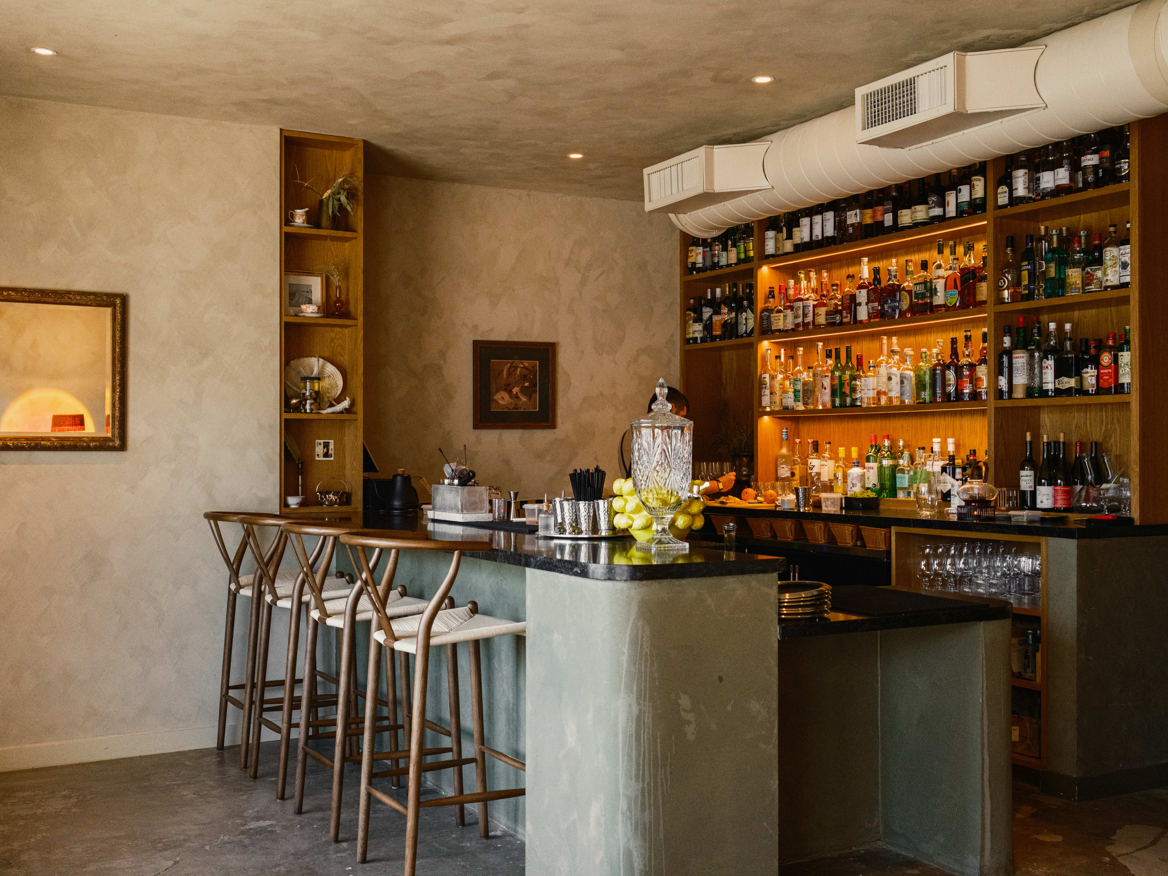 cocktail bar at Baso with a small bar top, four chairs, and an earthy/moody color palette with a back wall full of bottles