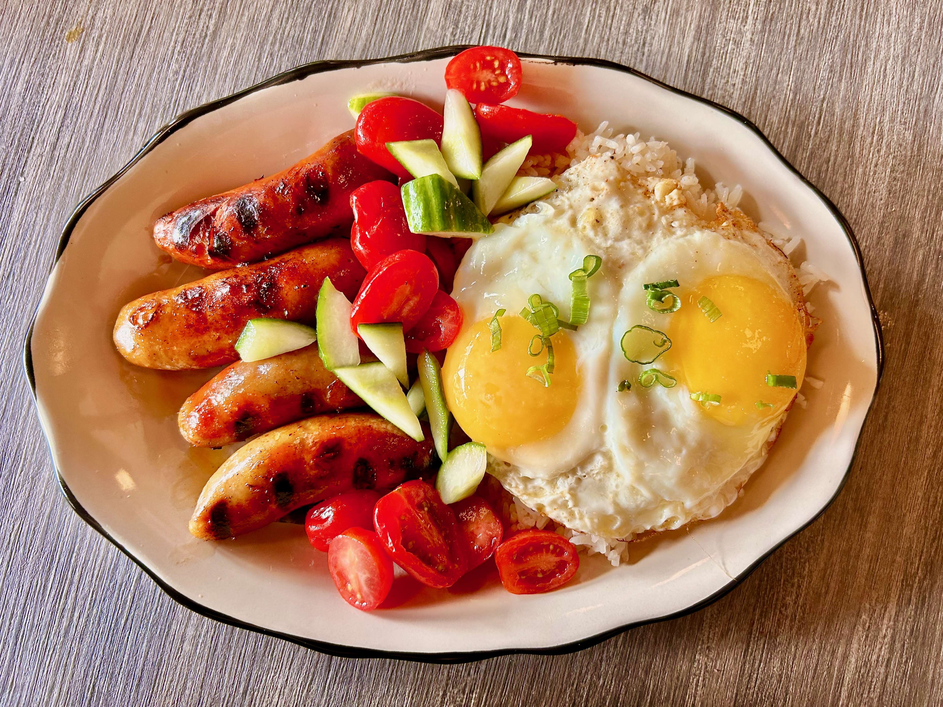 A plate of longanisa with garlic rice topped with eggs and some tomatoes and cucumber.