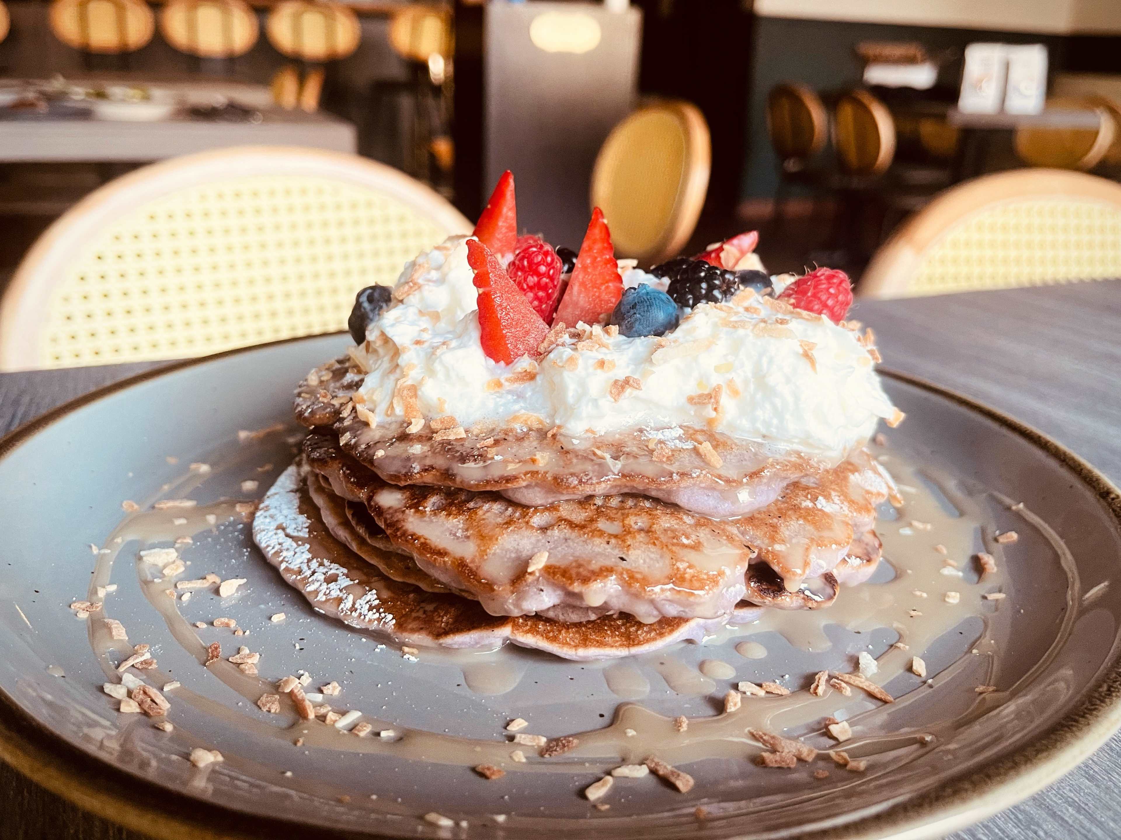 Stack of ube pancakes on a plate topped with whipped cream, fruit, and shredded coconut