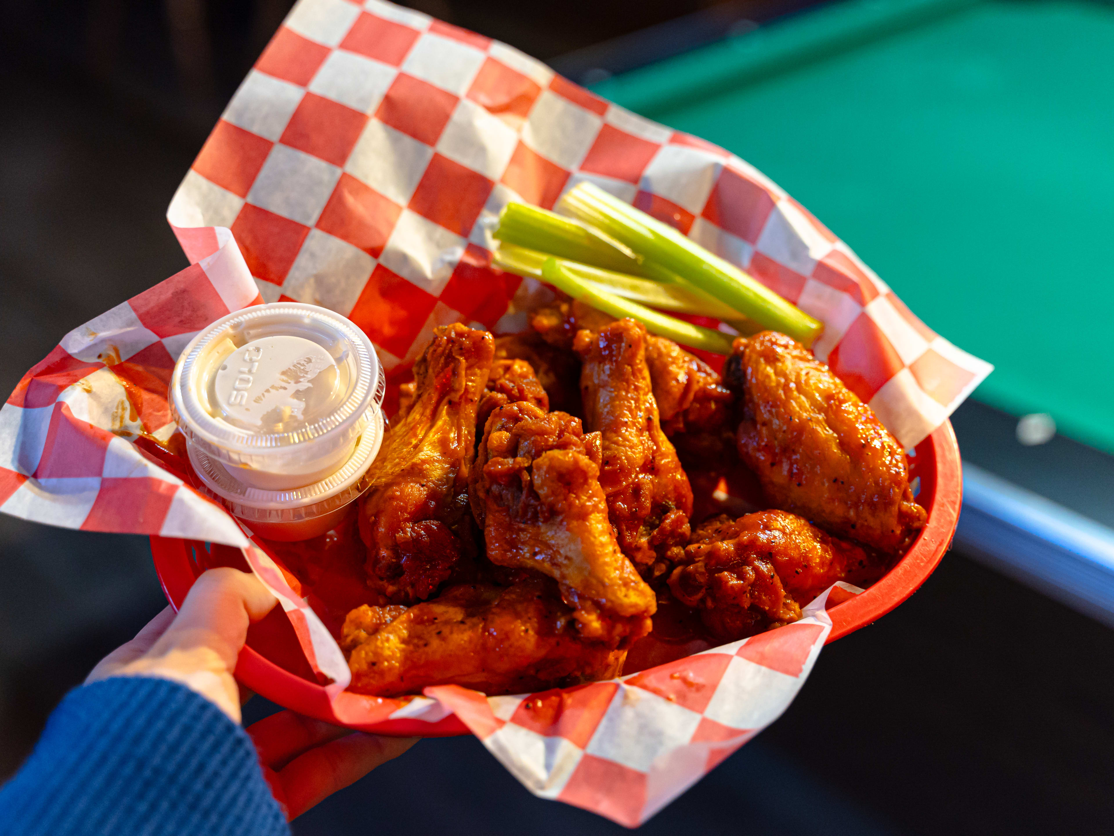 Chicken wings from Bird's Nest in a red basket with dipping sauce and celery held up in front of a pool table