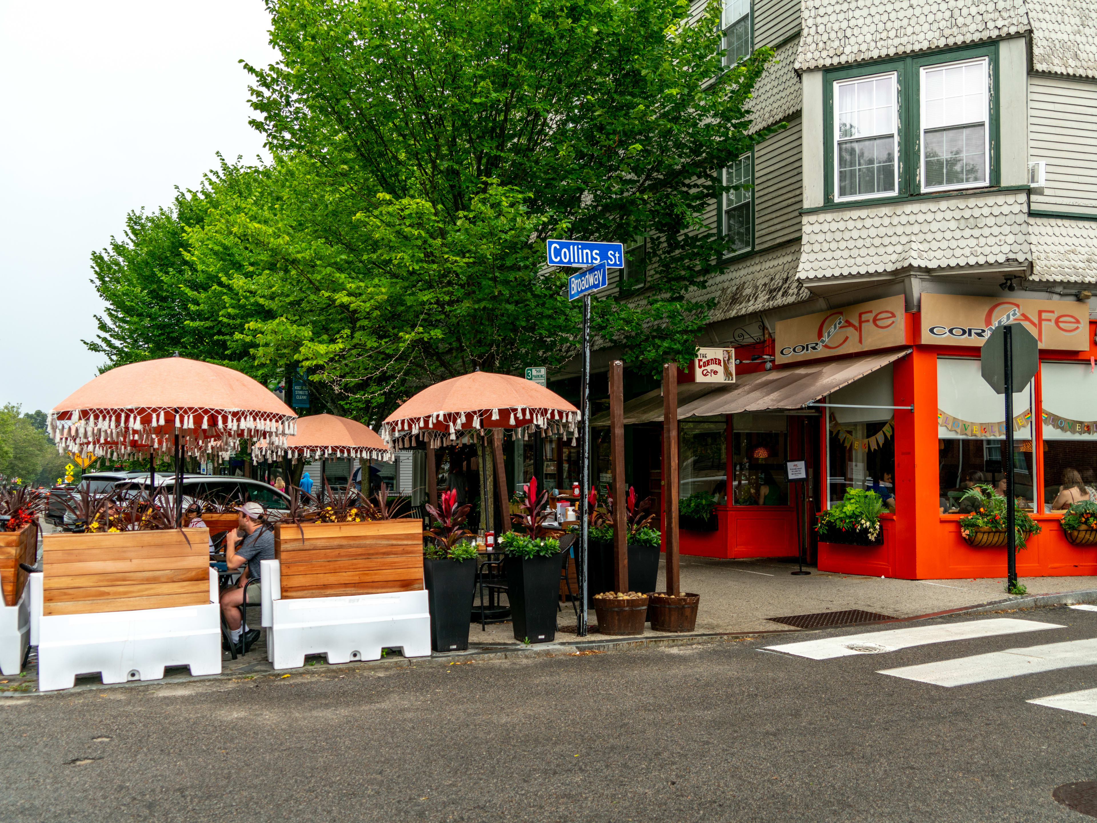 Corner Cafe exterior with window baskets filled with plants and an outdoor dining set up in the street with umbrellas and wooden walls