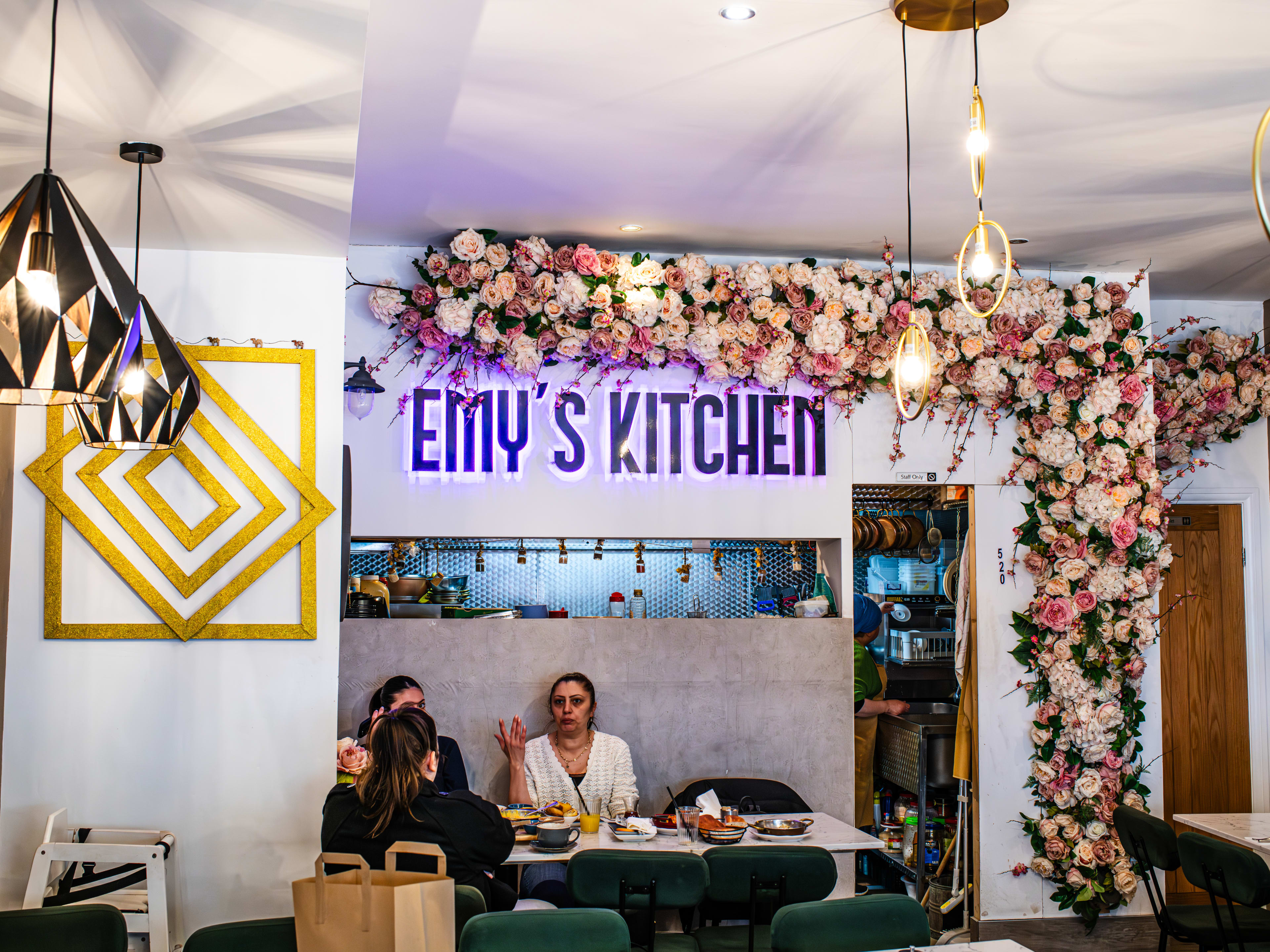 A bright dining room with fake flower garlands around one wall and a purple neon sign saying 'Emy's Kitchen'.