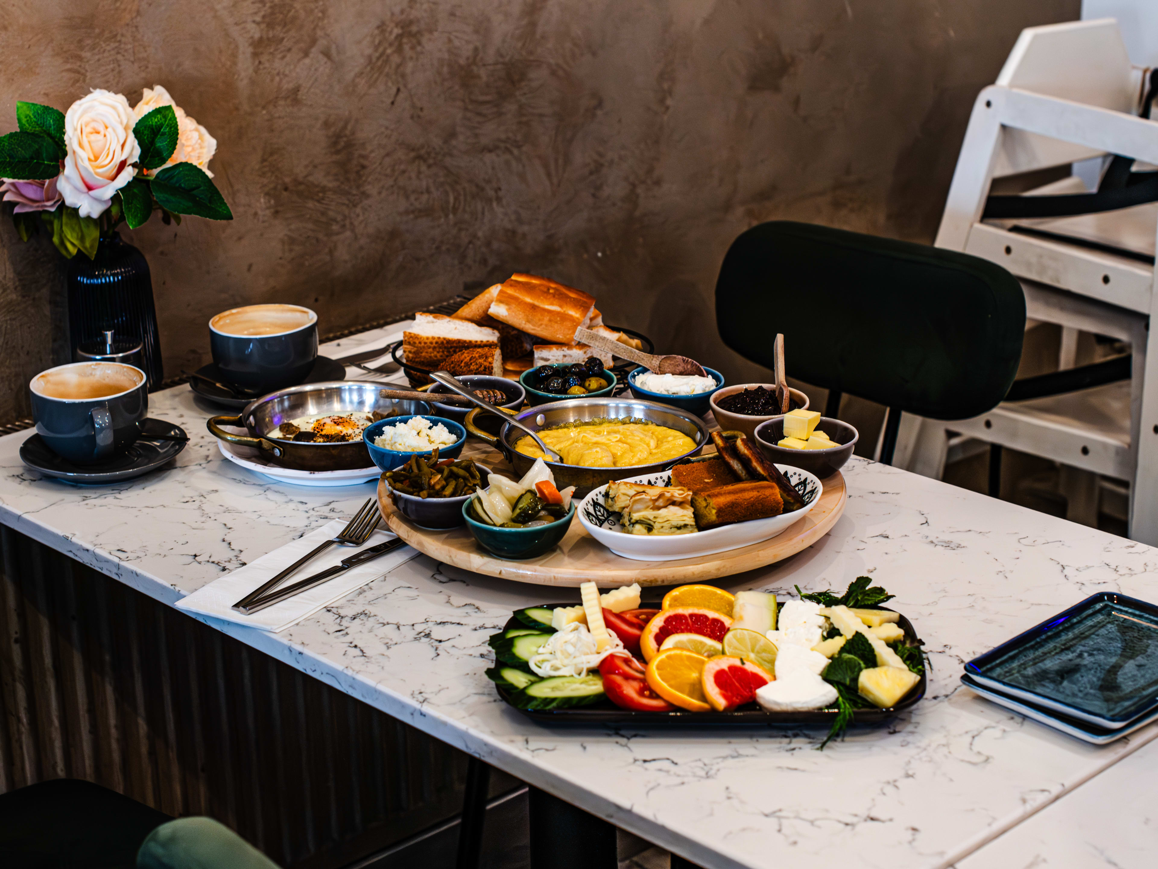 A pulled-back shot of a Turkish breakfast spread—with fruit, dips, coffees, cheese, and bread—on a marble-look table.