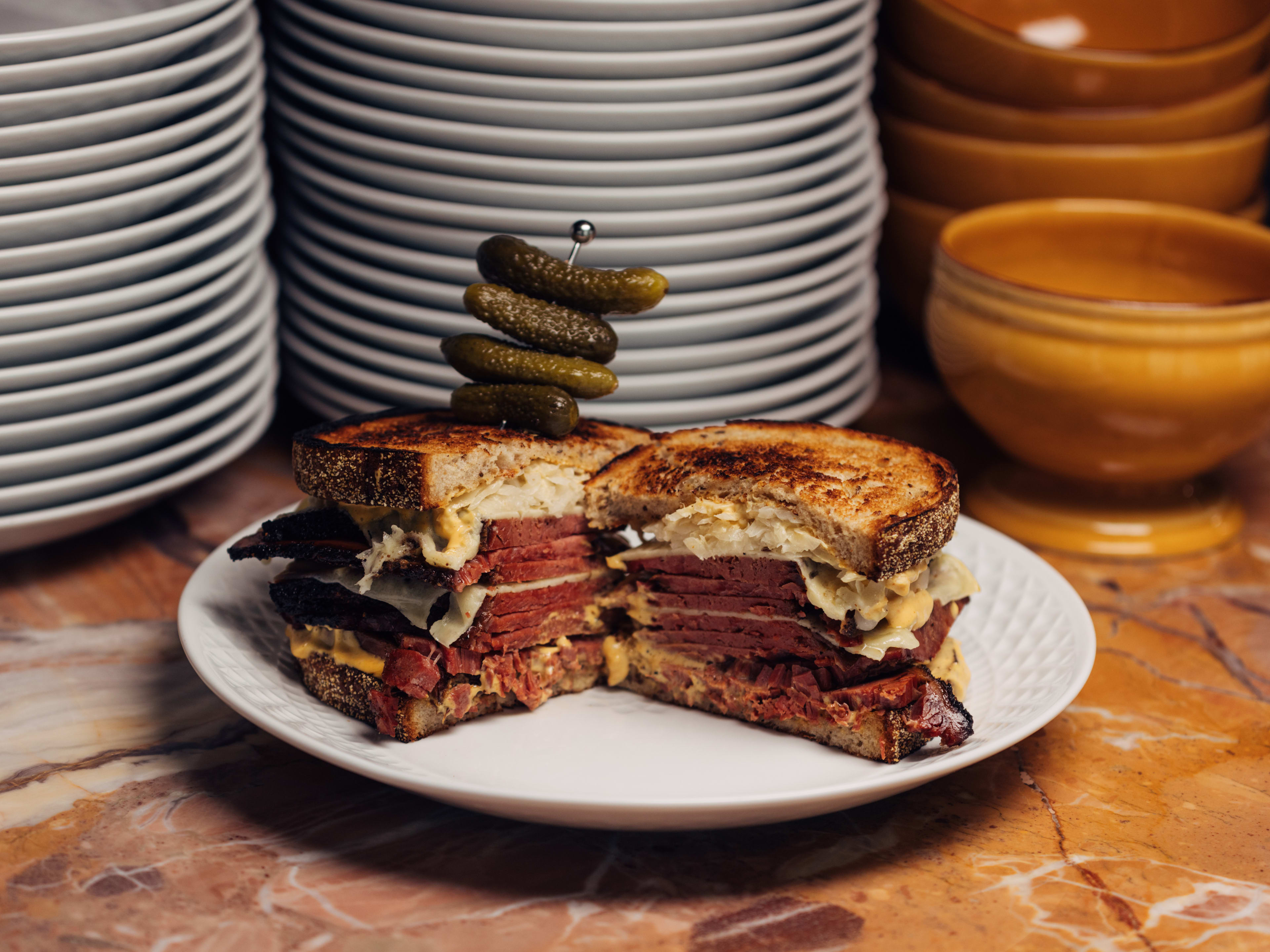 A pastrami sandwich cut in half with a skewer of cornichons in front of a stack of plates and bowls