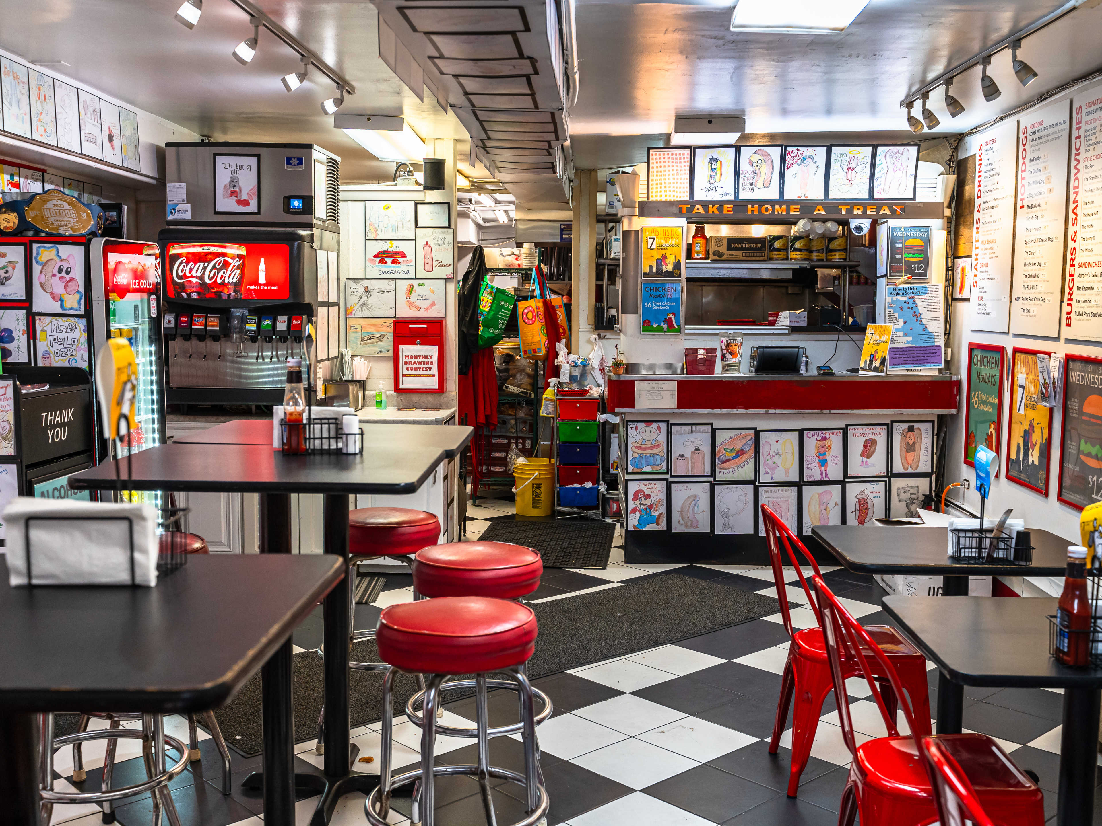 Low-ceilinged room with checkered floors, red bar stools at high tops, and hot dog wall art