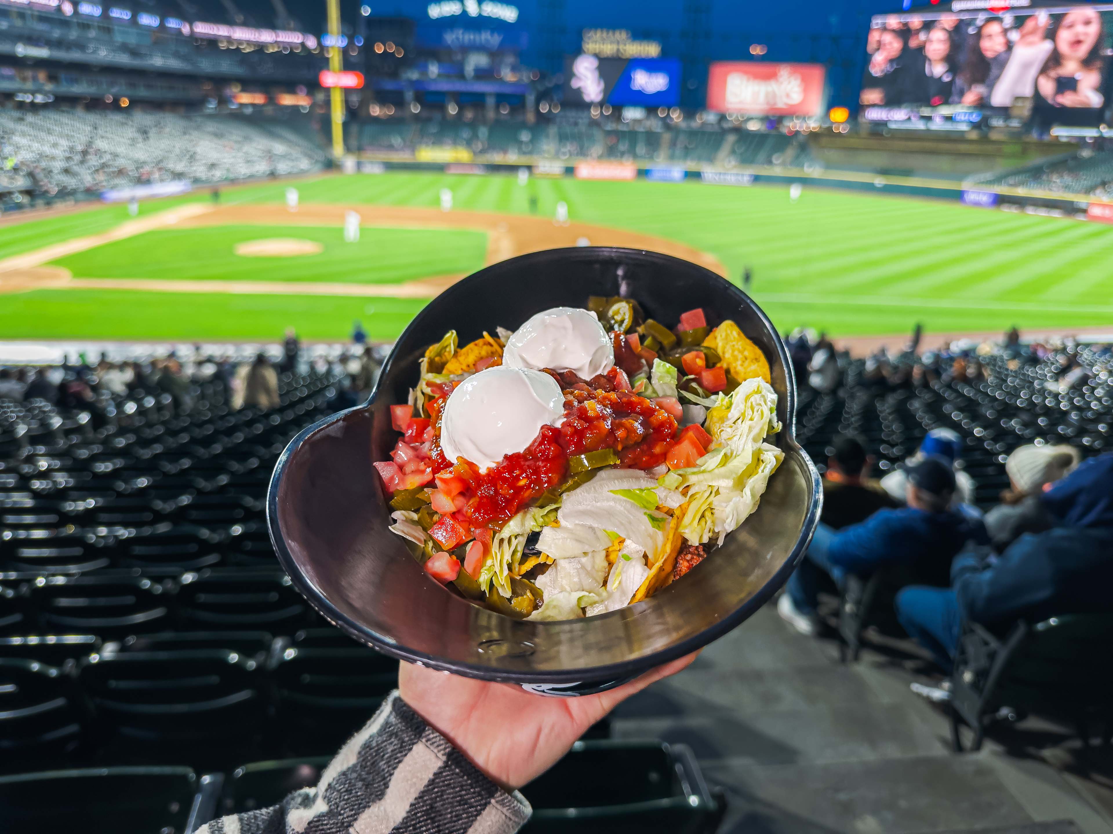 Massive souvenir helmet filled with nachos held up in front of a baseball diamond and stadium seating