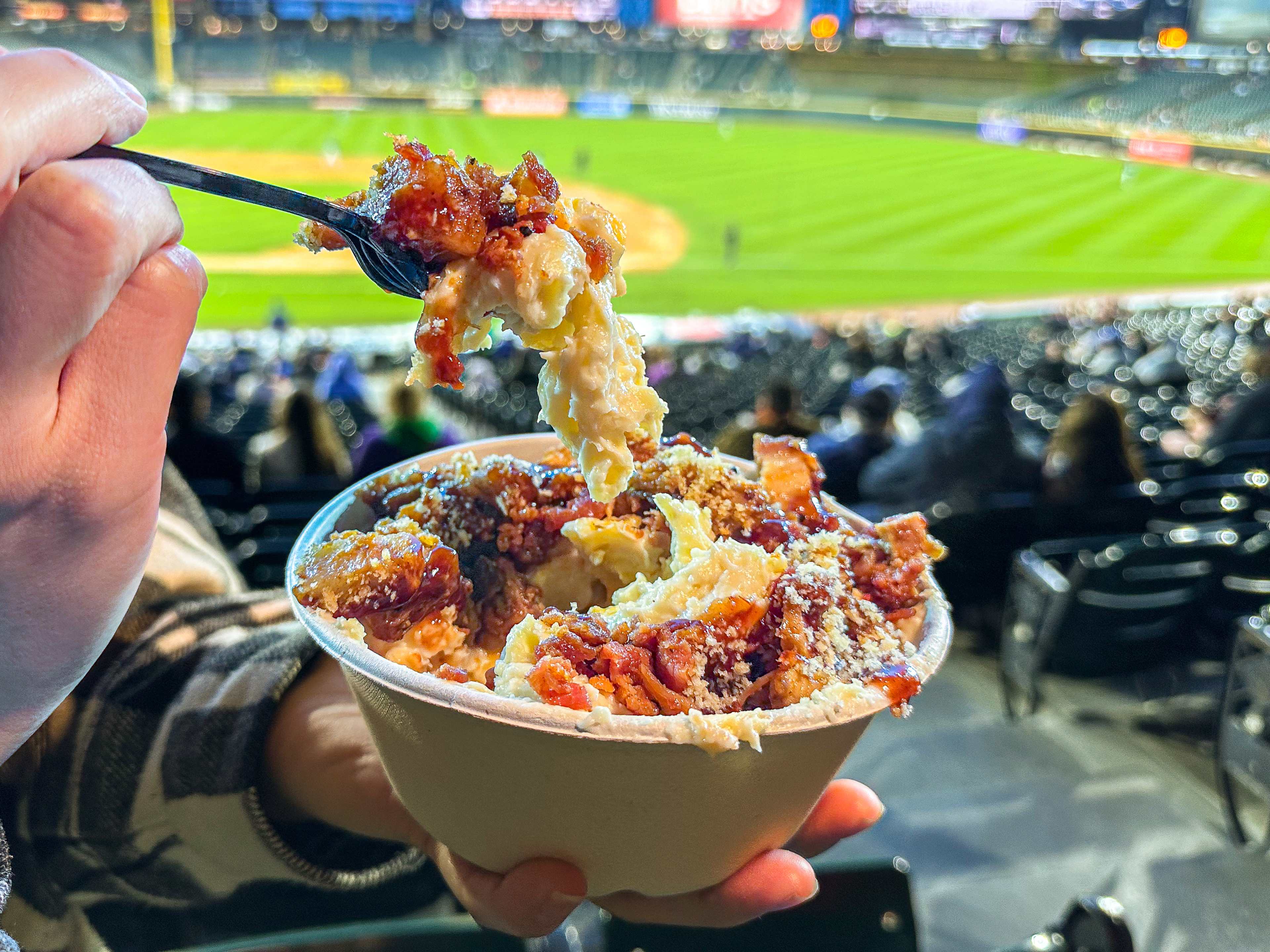 Bowl of macaroni and cheese with pieces of pork belly and bacon held up in front of a baseball diamond