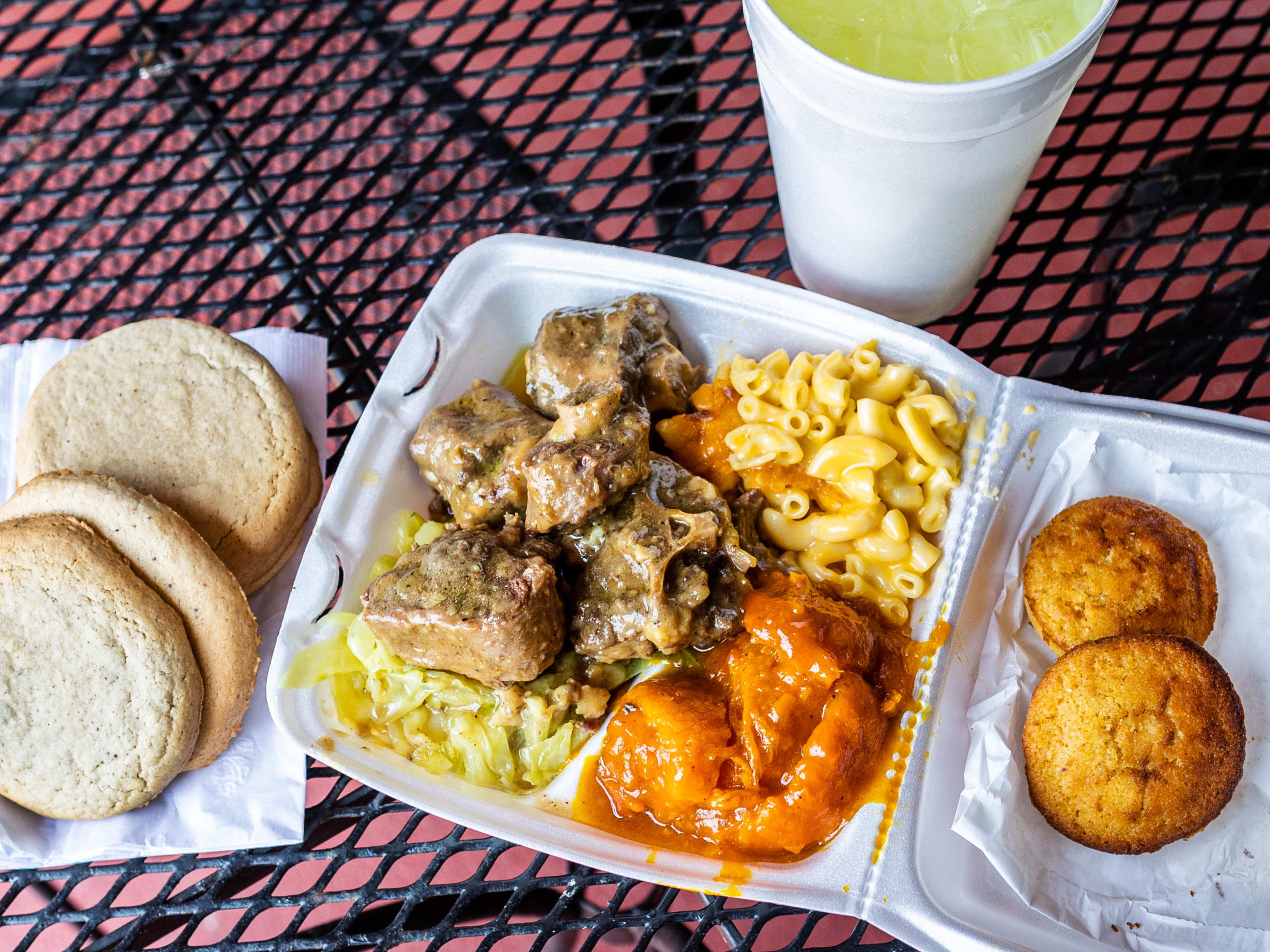 A takeaway styrofoam container of cornbread muffins, meats, mac and cheese sides, and three sugar cookies from Houston This Is It! Soul Food. A lemonade is in a styrofoam cup.