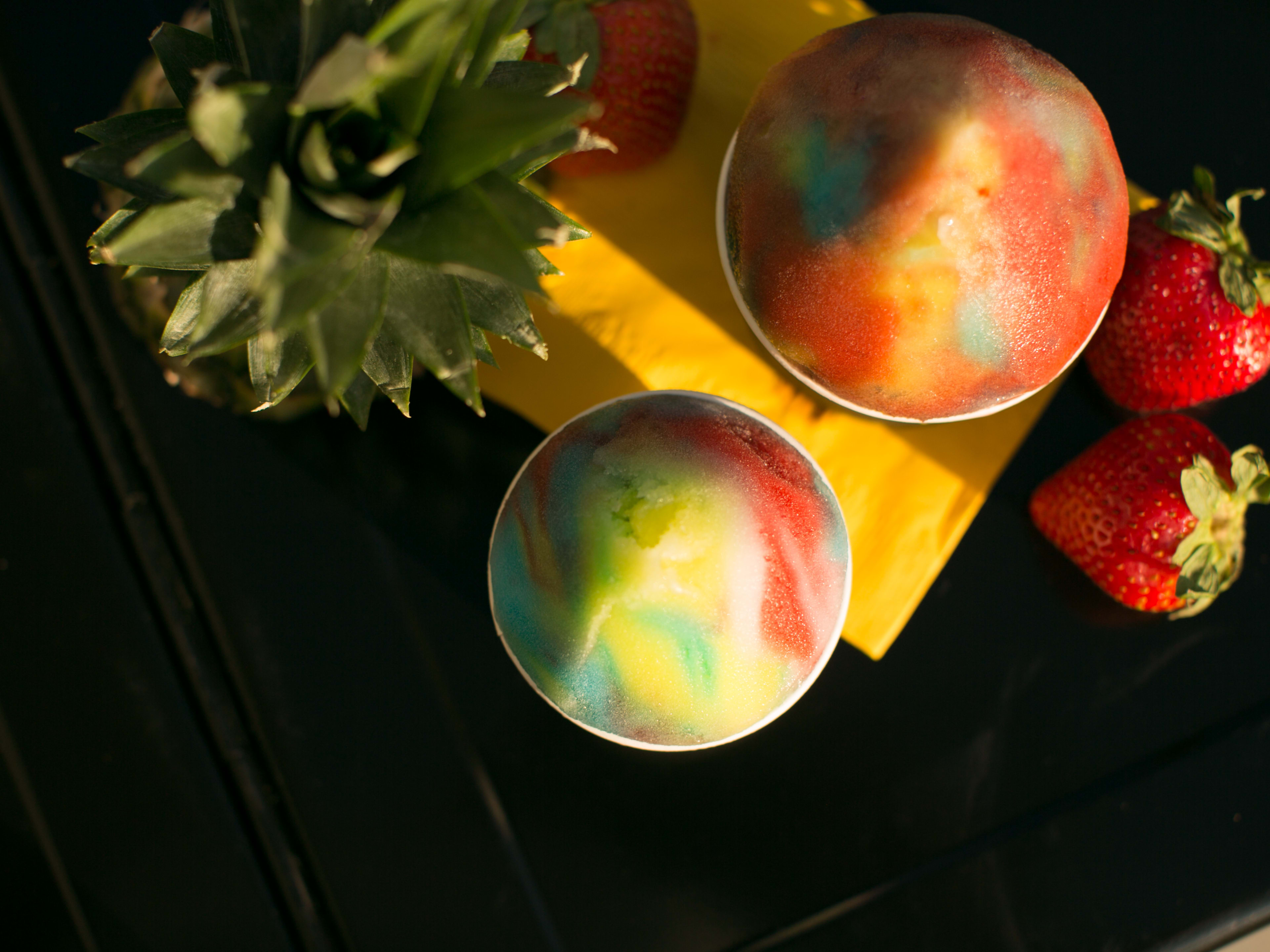 Frozen ice with swirls of brights colors on a black table next to stawberries and pineapple.