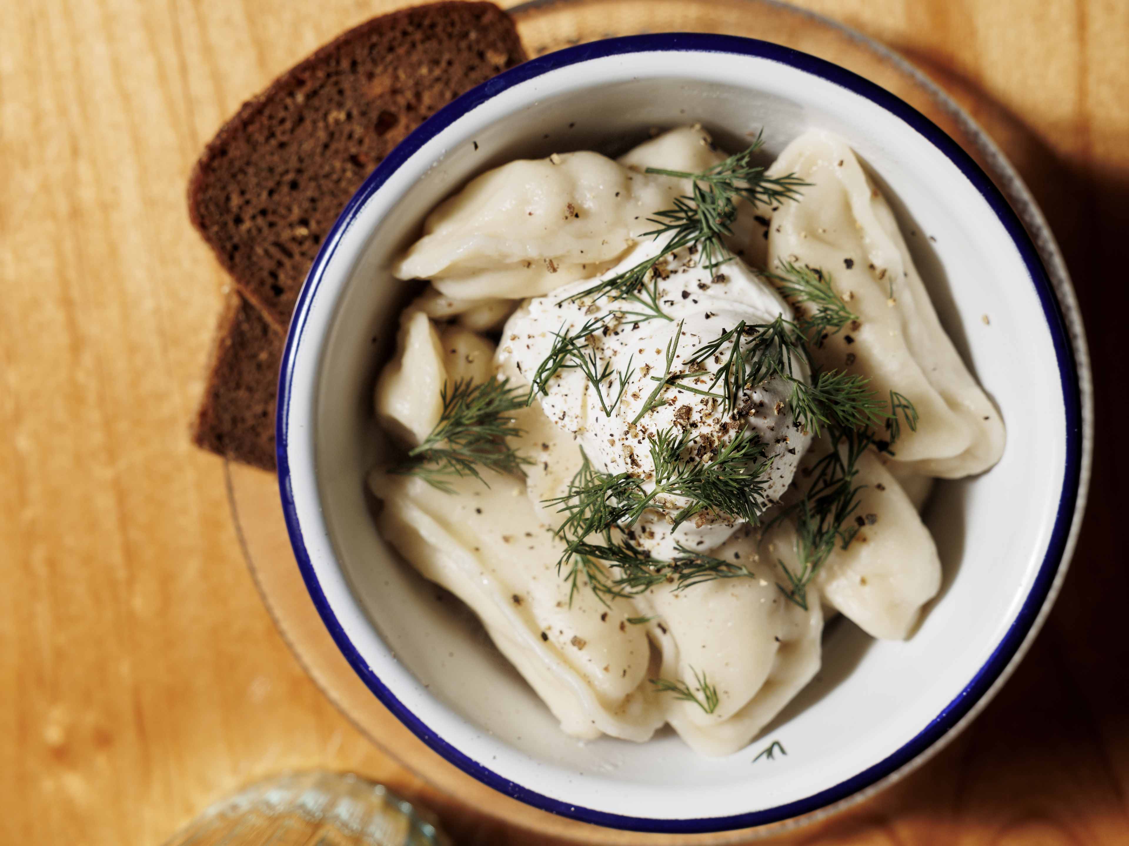 Bowl of pelmeni topped with sour cream and dill, alongside a slice of dark rye bread