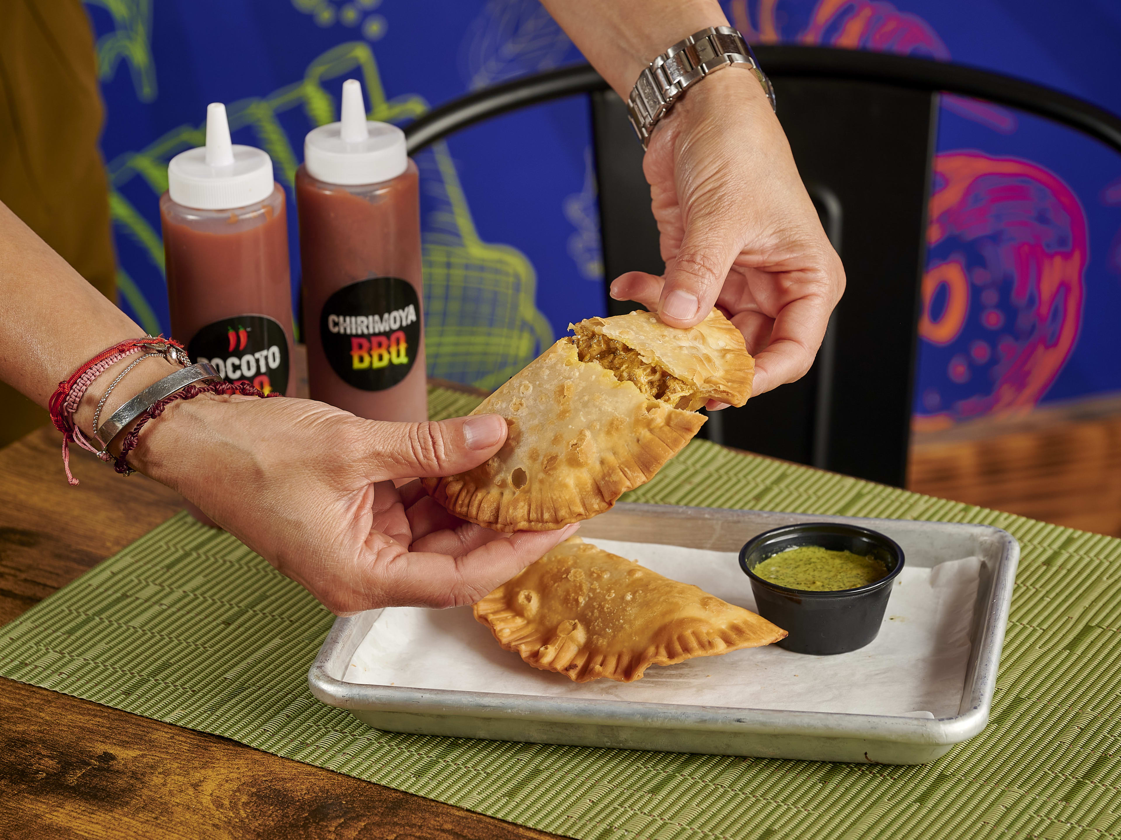 An empanada ripped open by a set of hands, green sauce, and two bottles of barbecue in the background