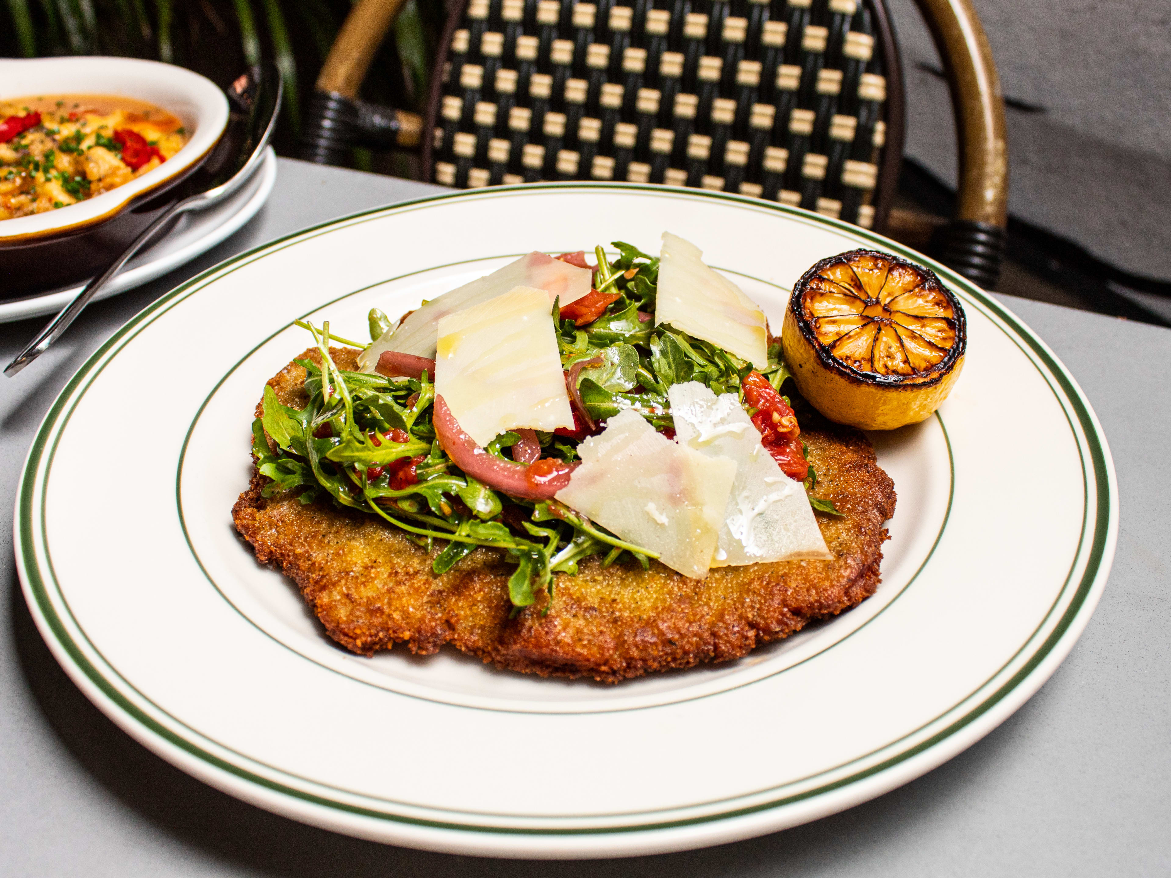 Breaded cutlet wtih arugula, tomatoes, shaved parm, and a burnt lemon half on a ceramic plate.