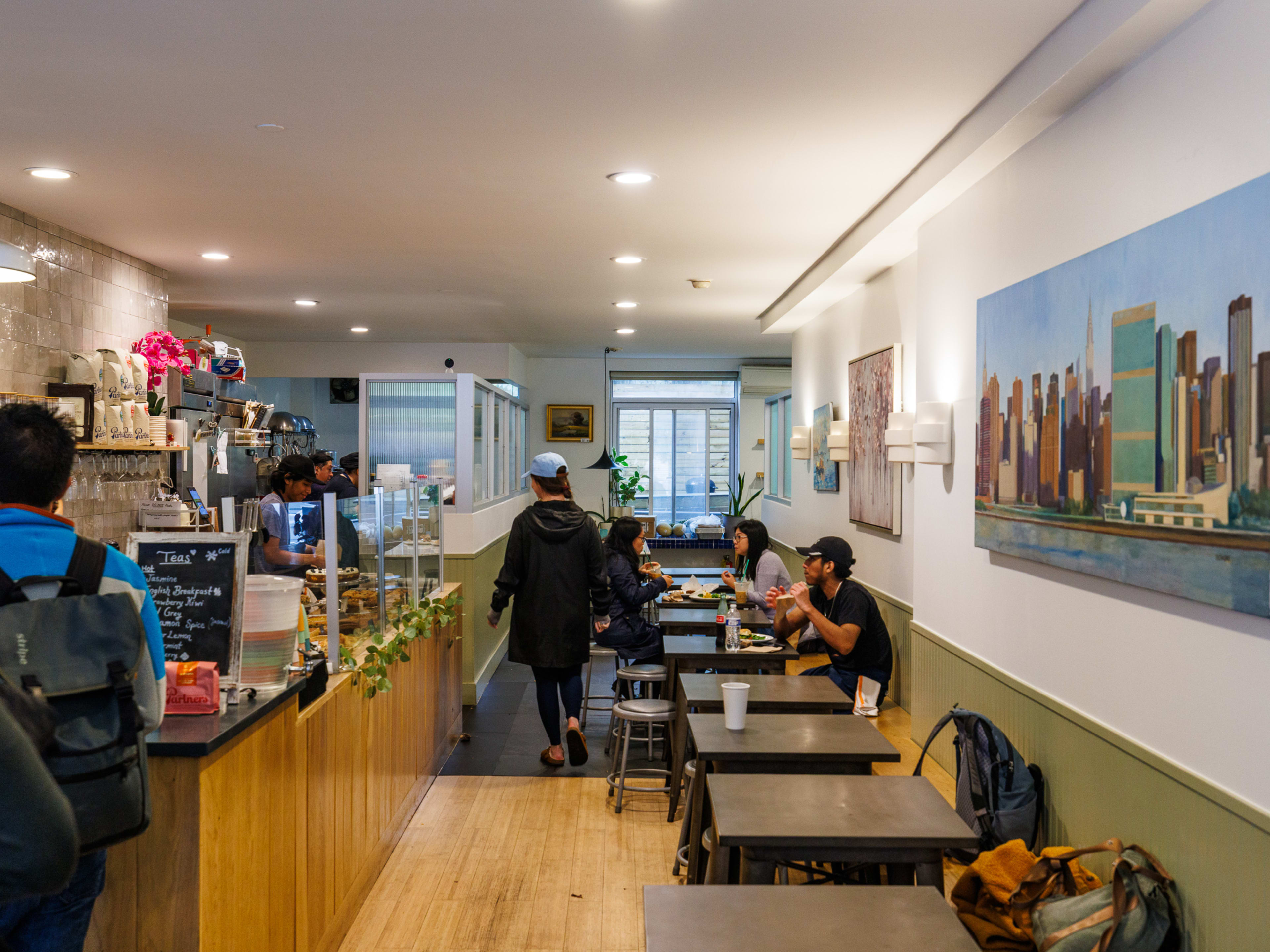 51st Bakery & Cafe interiors with people seated at wooden square two top tables on the right and customers waiting in line to order at the counter on the left