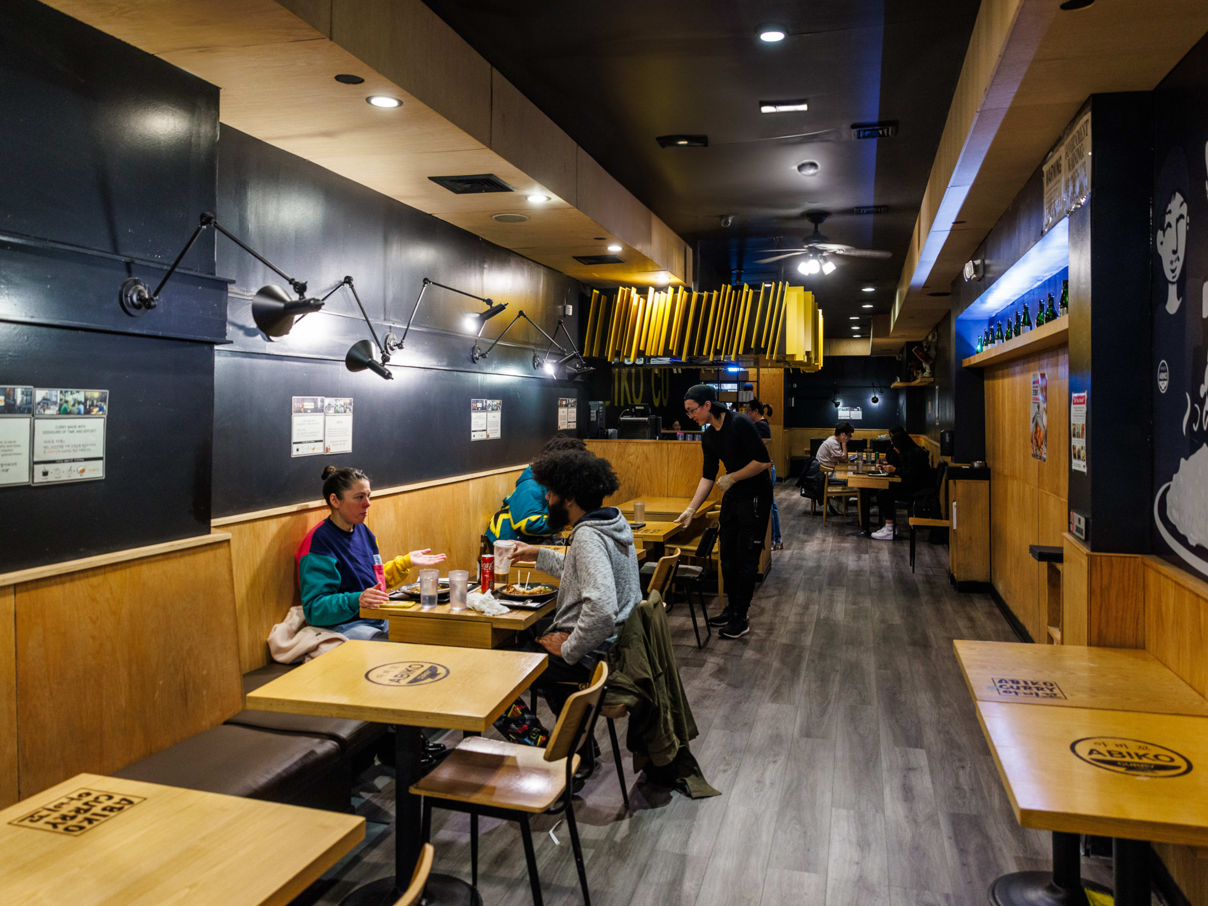 Abiko Cury interiors with black walls and people seated at wooden square tables