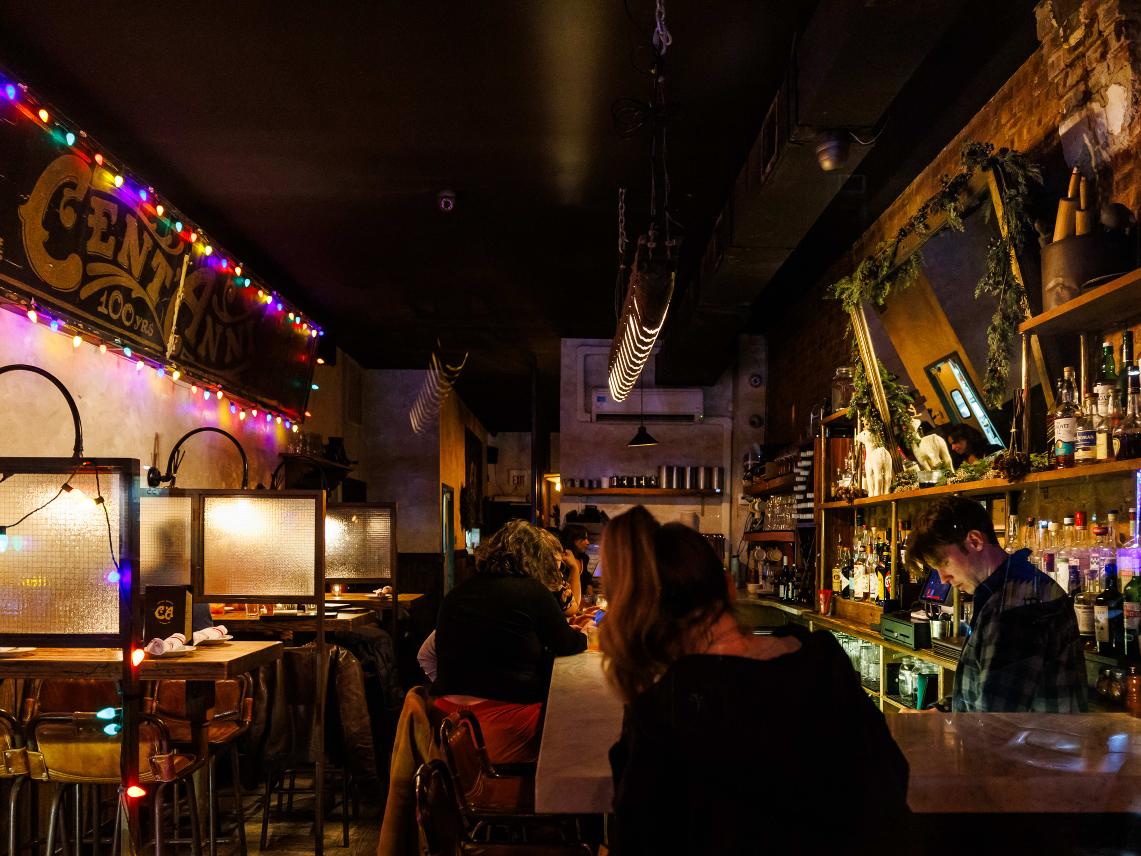 Cent'Anni interior with dim lighting, colorful string lights, high top seating with glass separators on the left, and people seated at the bar on the right