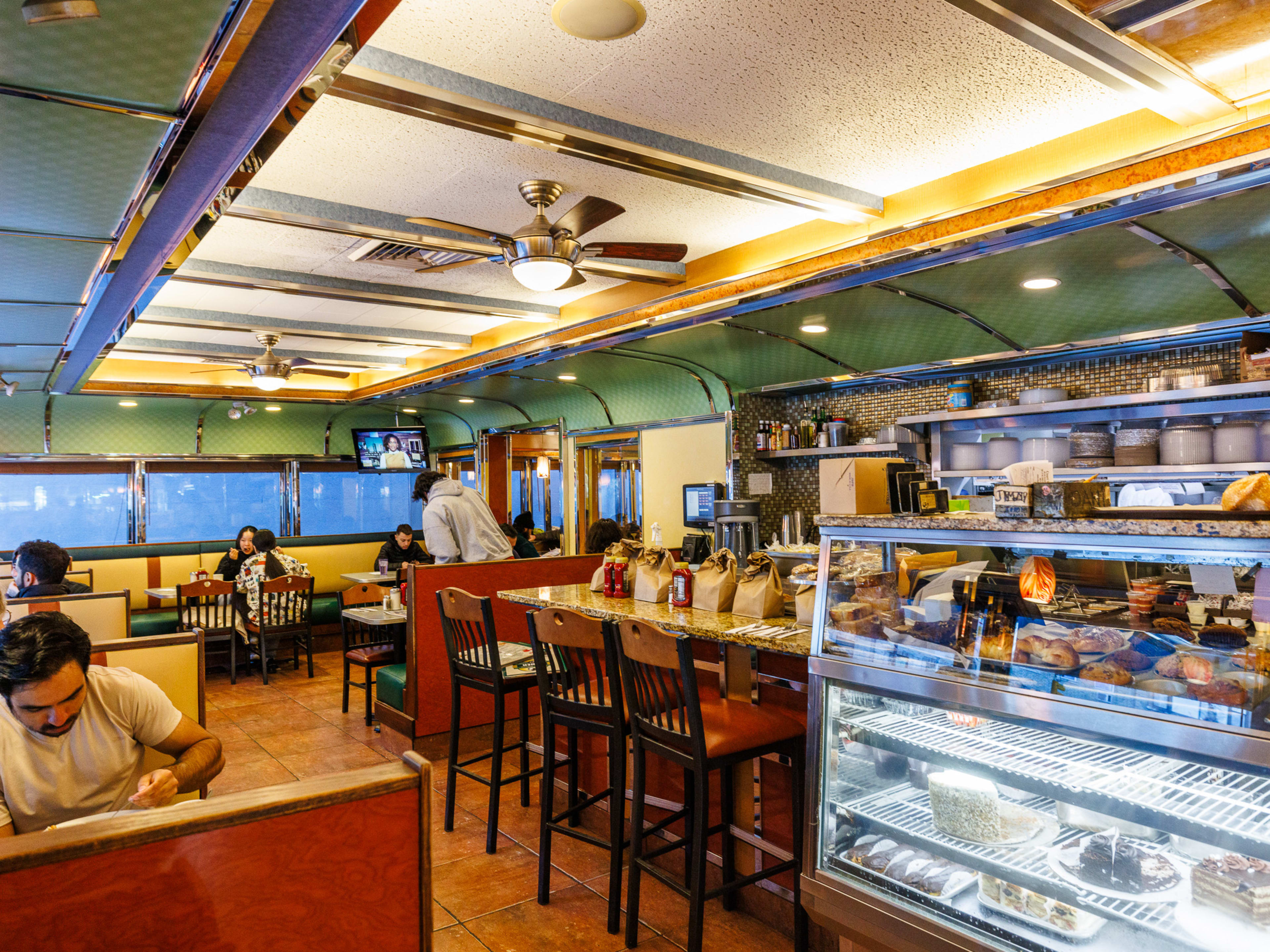 Court Square Diner interiors with rounded ceilings, cakes and pastries on display in the glass counter, and customers dining at yellow booth seating around the perimeter of the restaurant