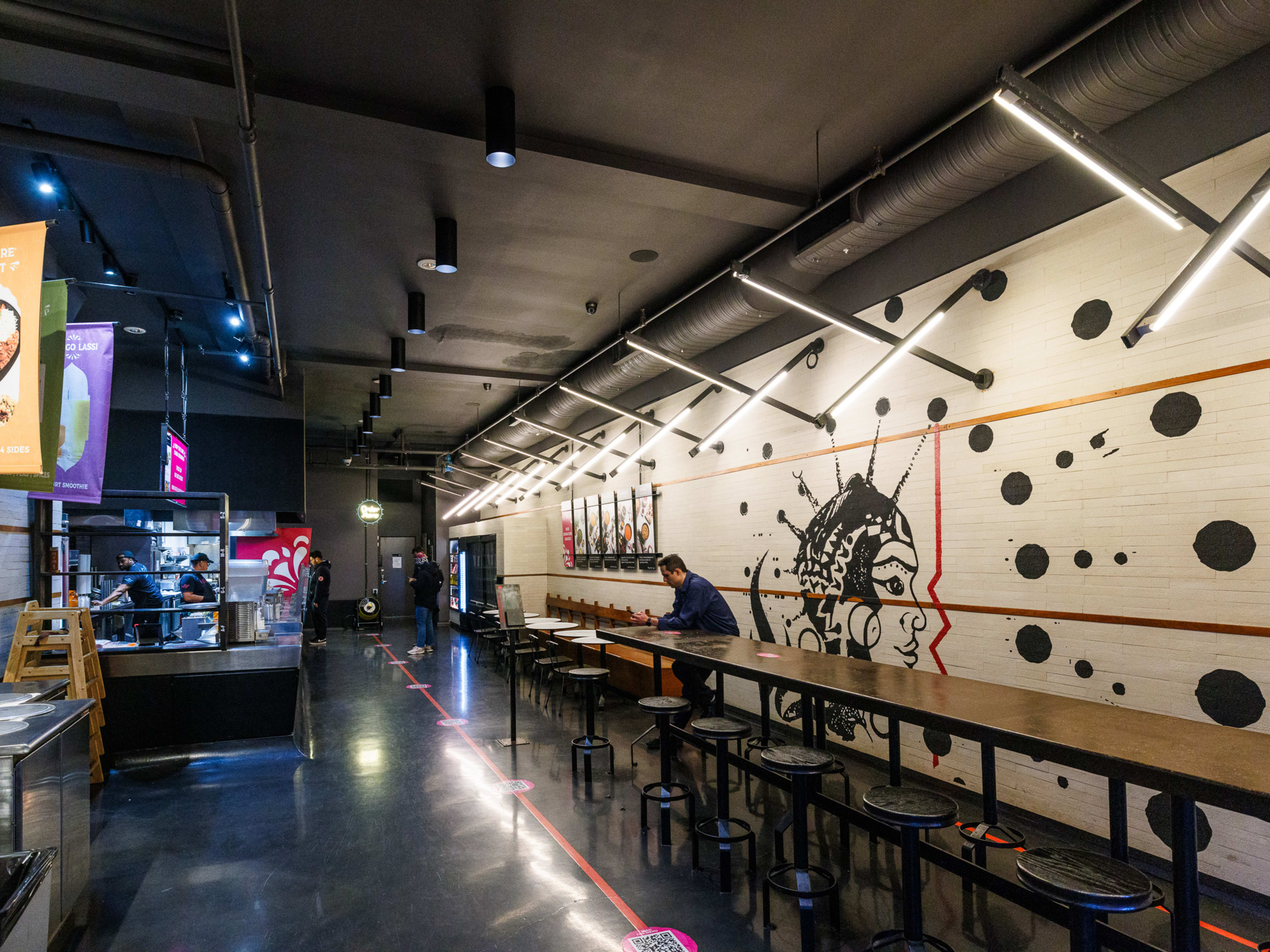 Deep Indian Kitchen interior with long counter seating with metal stools and a black and white painted mural on the wall