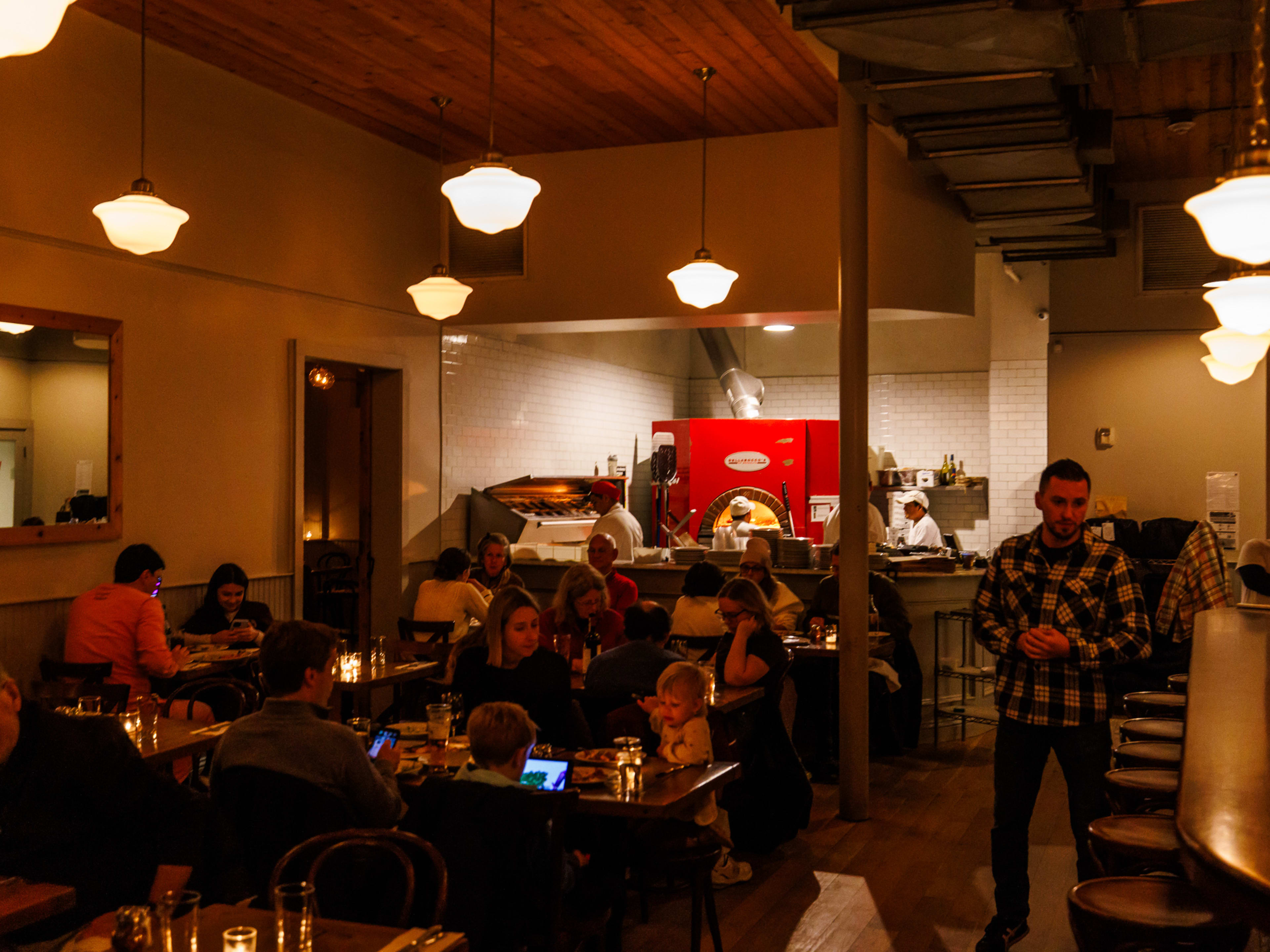 Dellarocco's interior with dim lighting, people dining at wood tables, and a pizza oven in the back