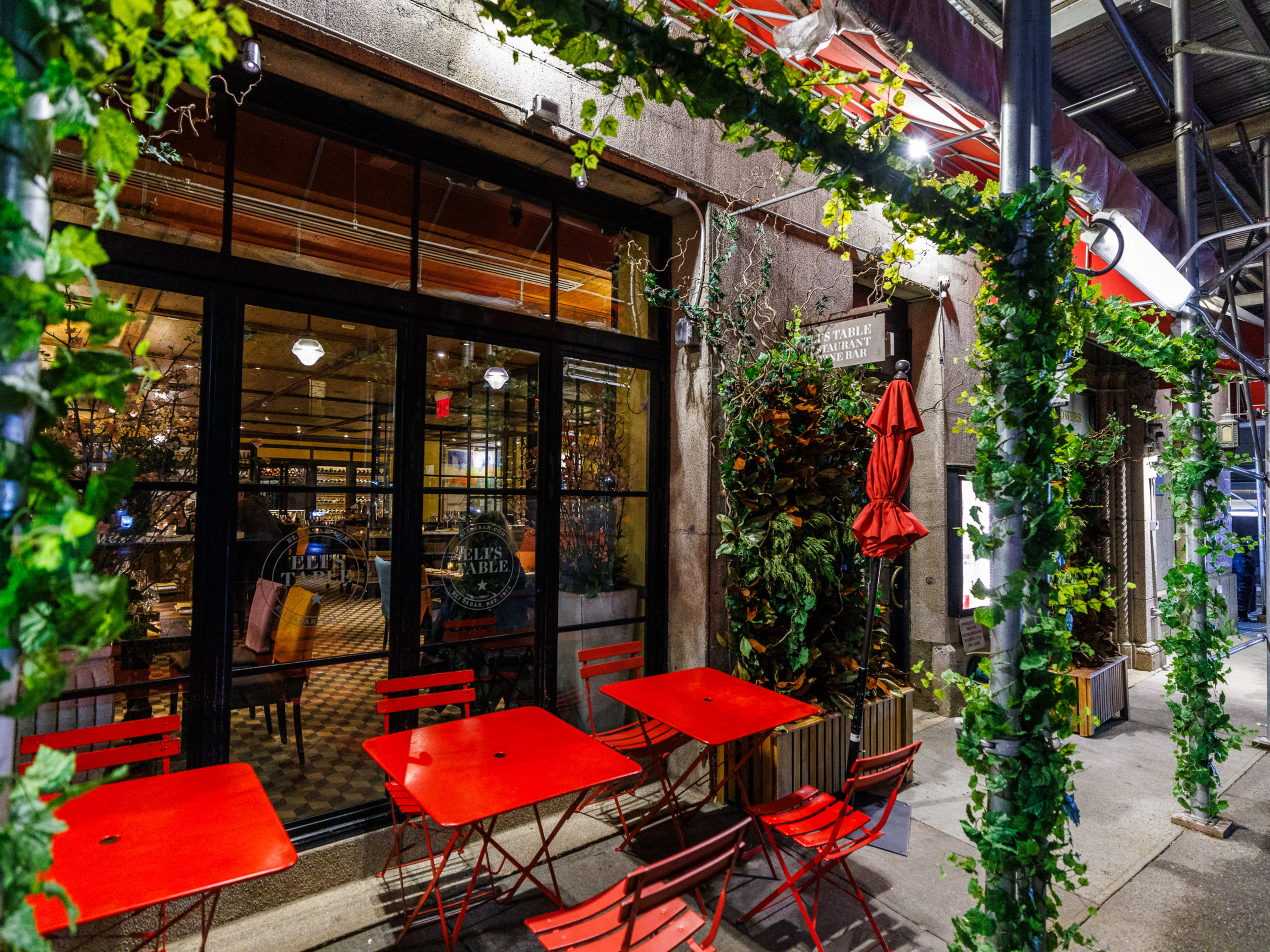 Eli's Table exterior covered in green vines and square red metal outdoor tables and chairs on the sidewalk