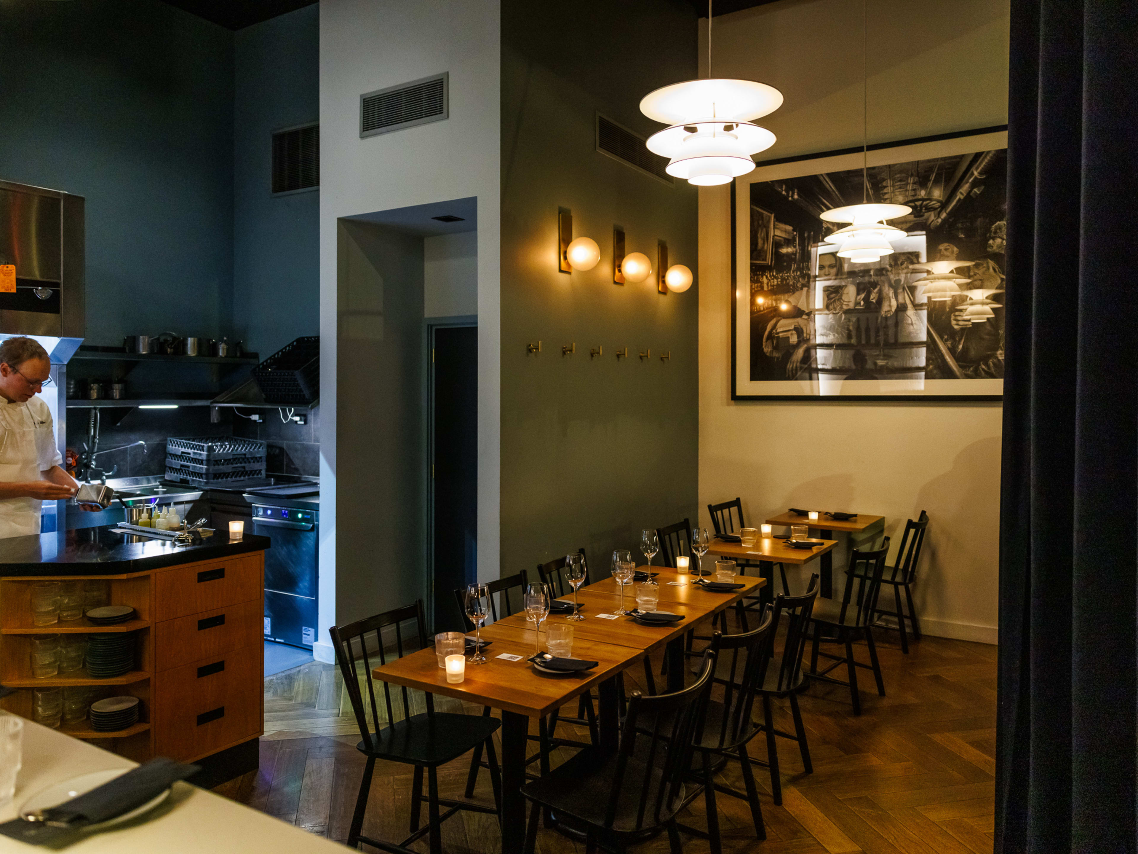 Farra interiors with a view into the kitchen on the left and multiple square wooden tables set for dinner on the right with candles and dim lighting