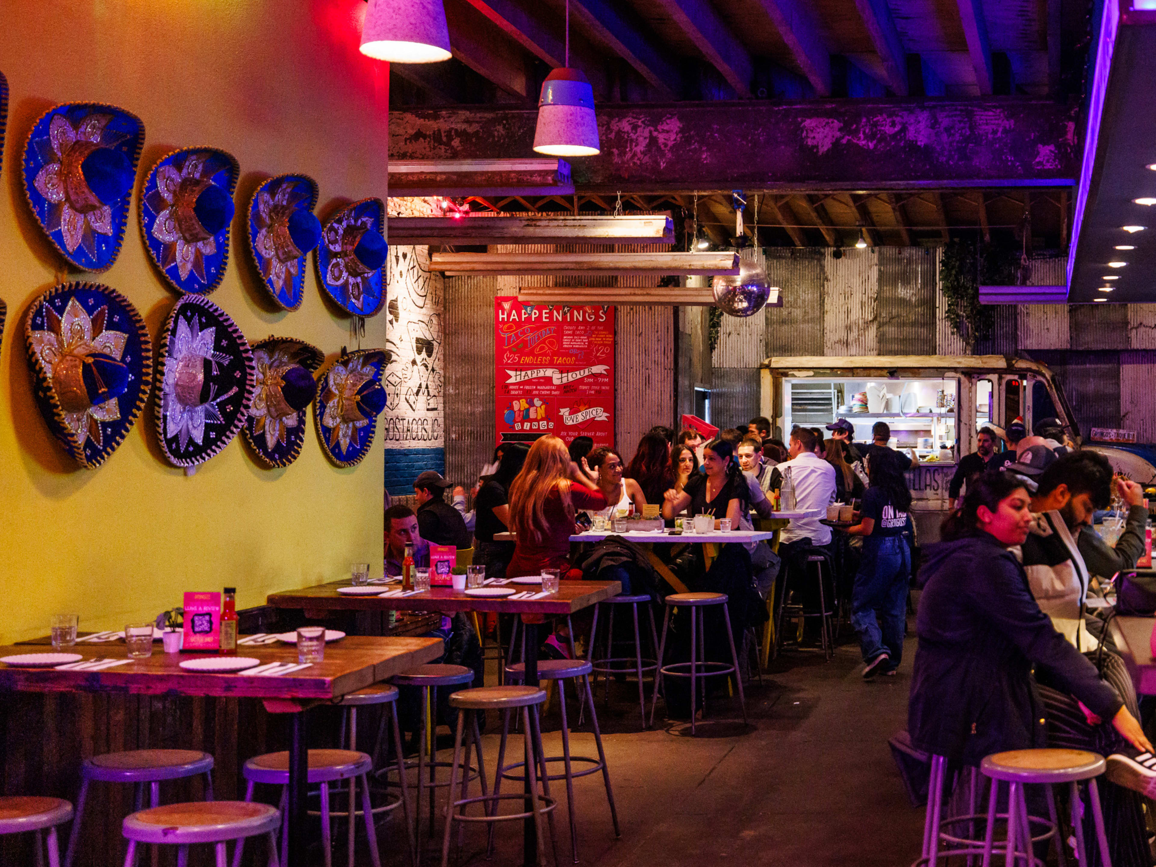 Gringo's interiors with sombreros hanging on the left wall, dim colorful lighting, people seated at high top tables, and a taco truck at the back of the restaurant.