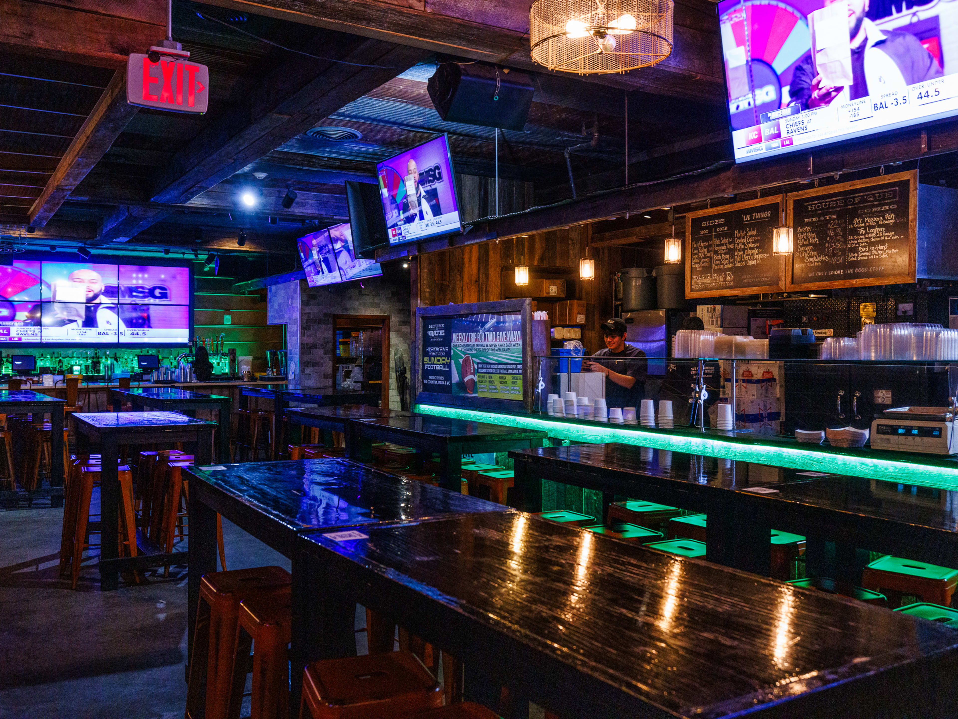 House of Que interiors with dim lighting, multiple tvs hanging from the ceiling, and long wooden communal high top tables