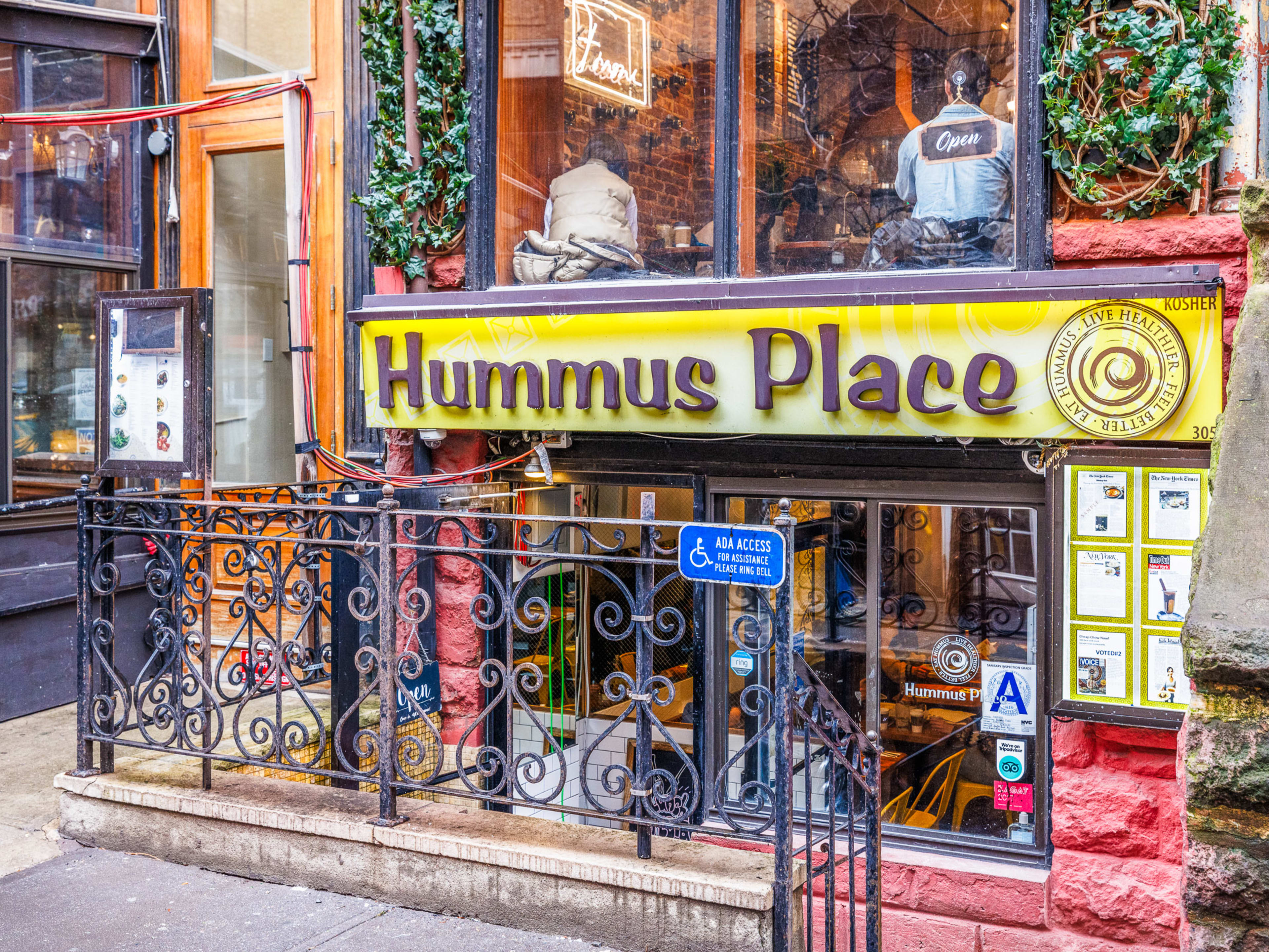 Hummus Place exterior with yellow restaurant sign and large windows with a view into the restaurant