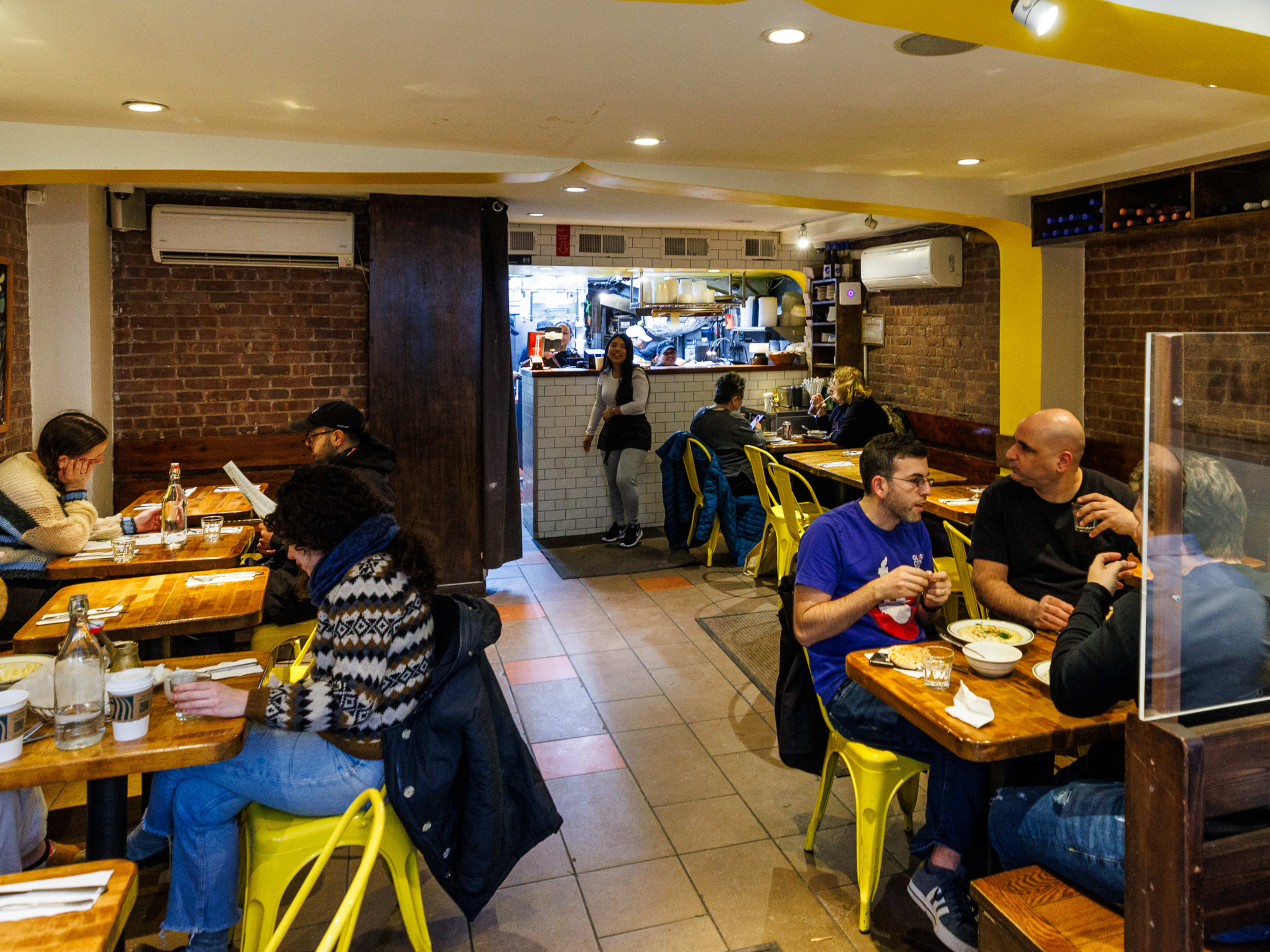 Hummus Place interiors with brick walls and customers seated at square wooden tables with yellow metal chairs