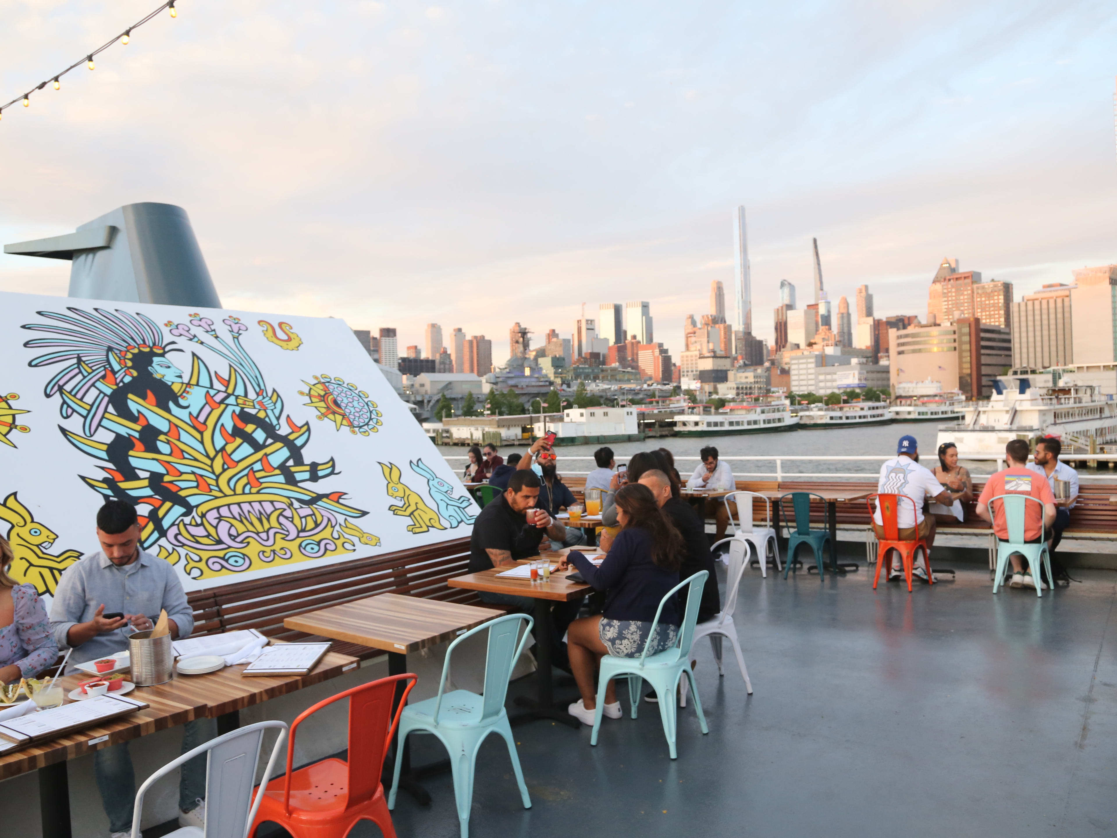 La Barca Cantina boat  with people seated at wooden tables and views of the manhattan skyline in the background at sunset