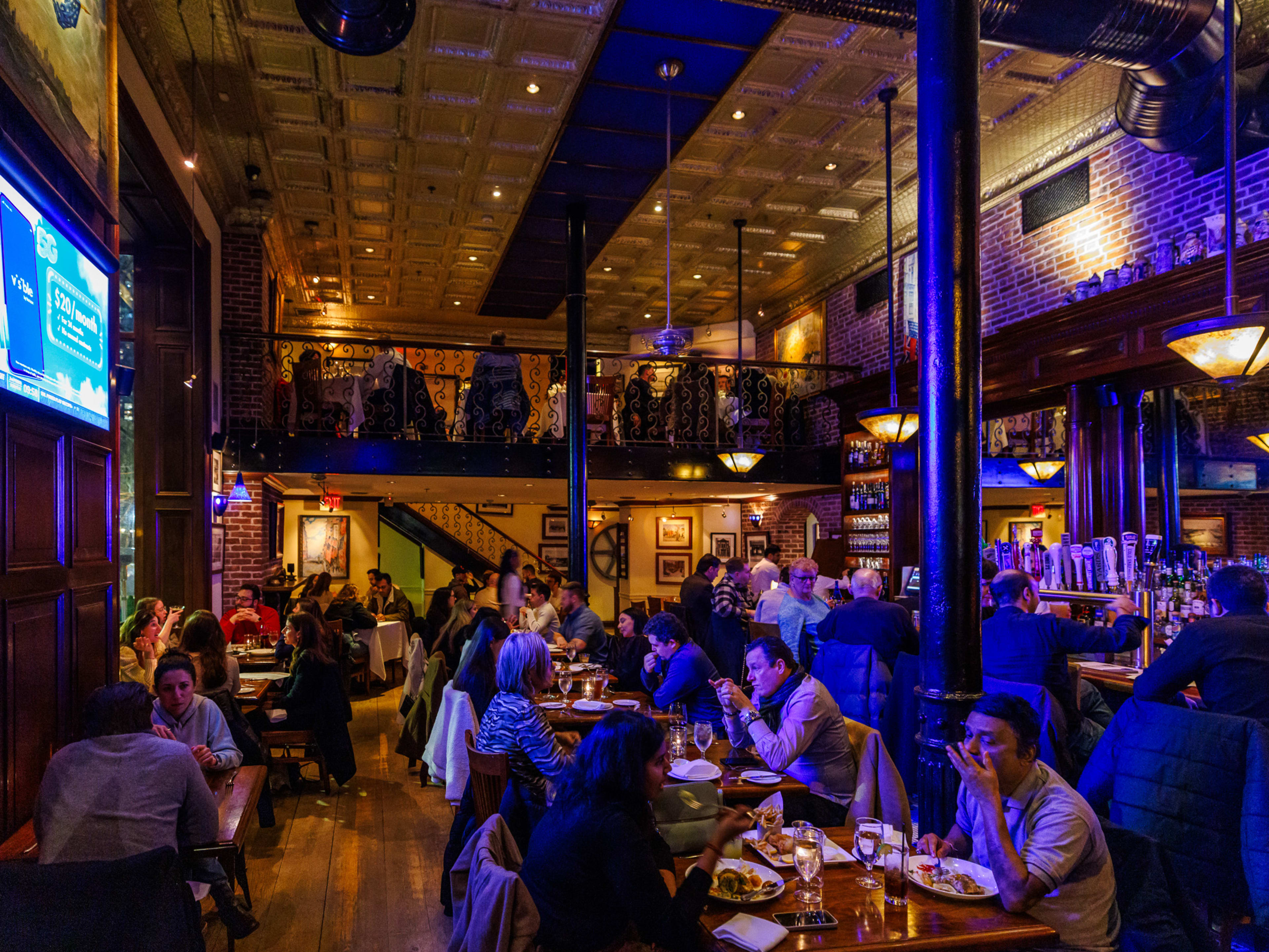 Light Horse Tavern interiors with blue dim lighting, people seated at wooden tables and at the bar, a balcony with additional seating in the back, and a large tv screen on the left wall.