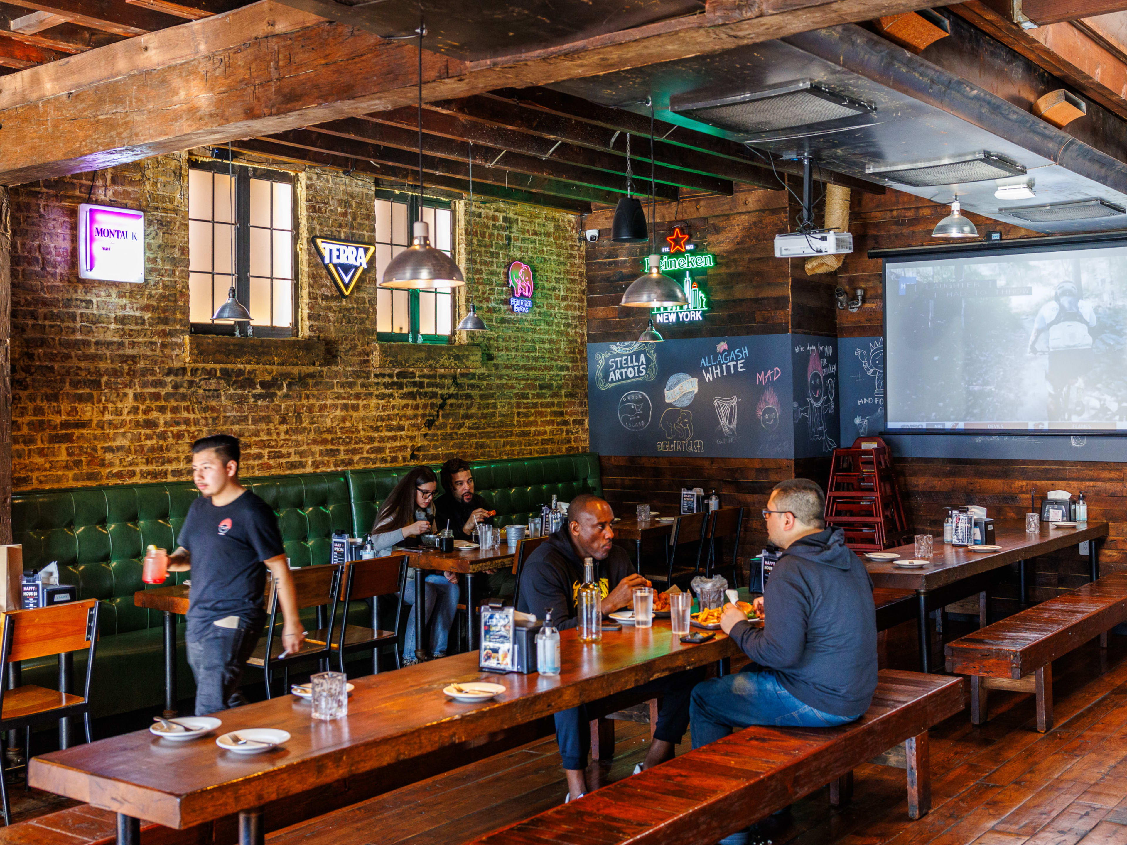 Mad For Chicken interiors with brick walls, people sitting at large wooden picnic tables, and a projection screen on the back wall