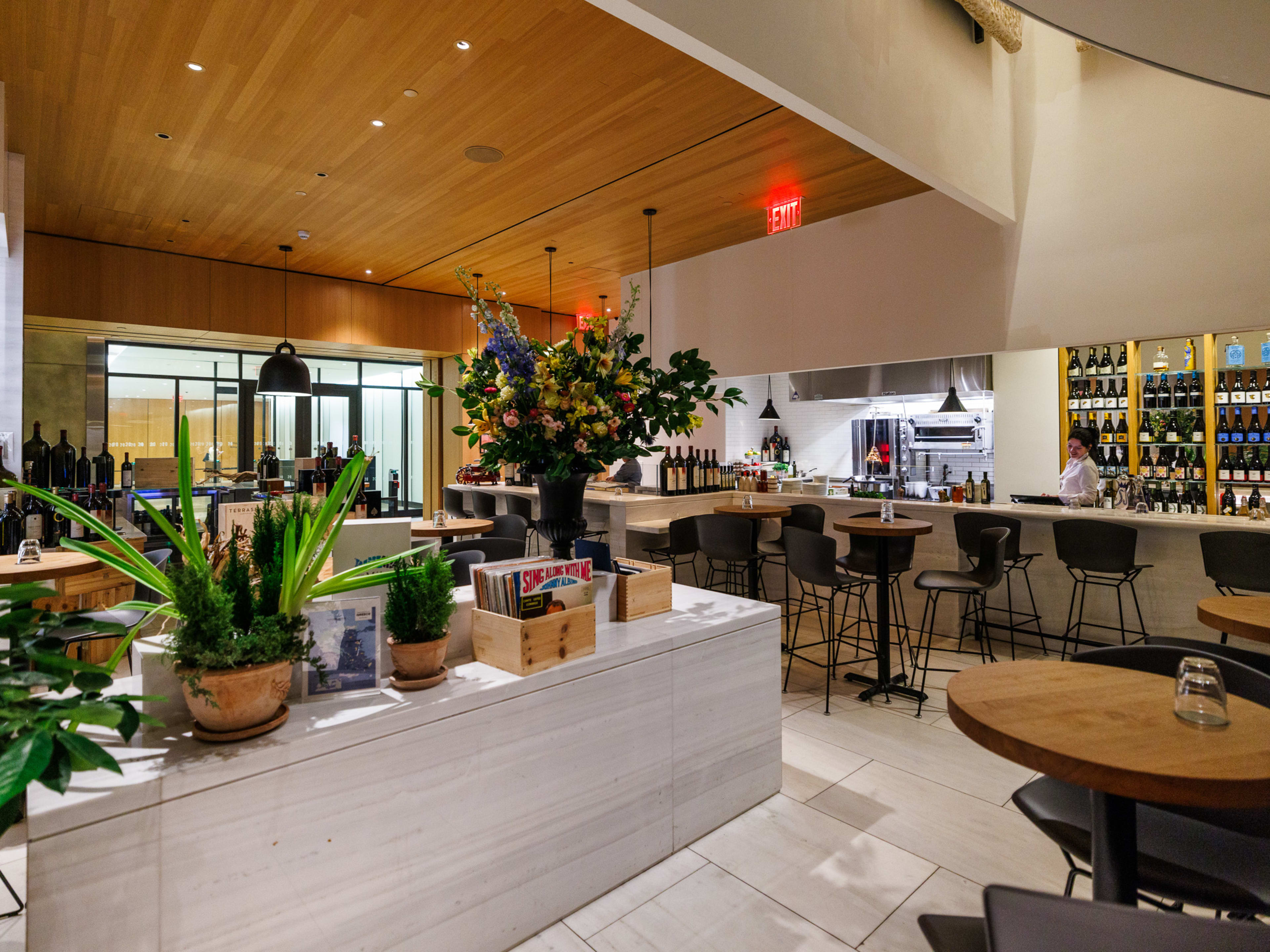 Milos Wine Bar interiors with high top seating, a wall filled with wine bottles behind the bar, and plants and flowers throughout the space