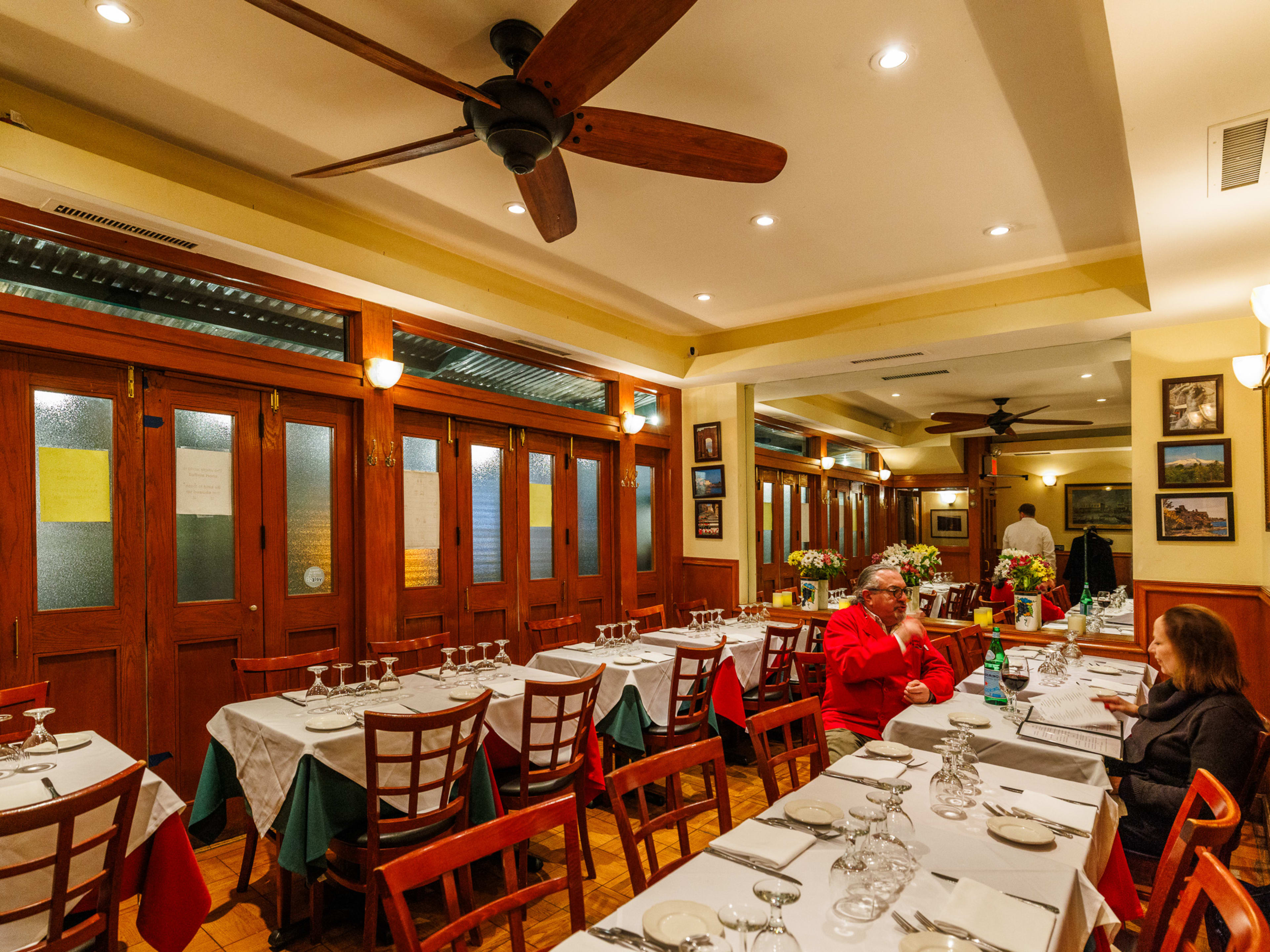 Nica Trattoria interiors with a large mirror on the back wall, wooden doors on the right wall, and multiple square tables with white table cloths and place settings on them