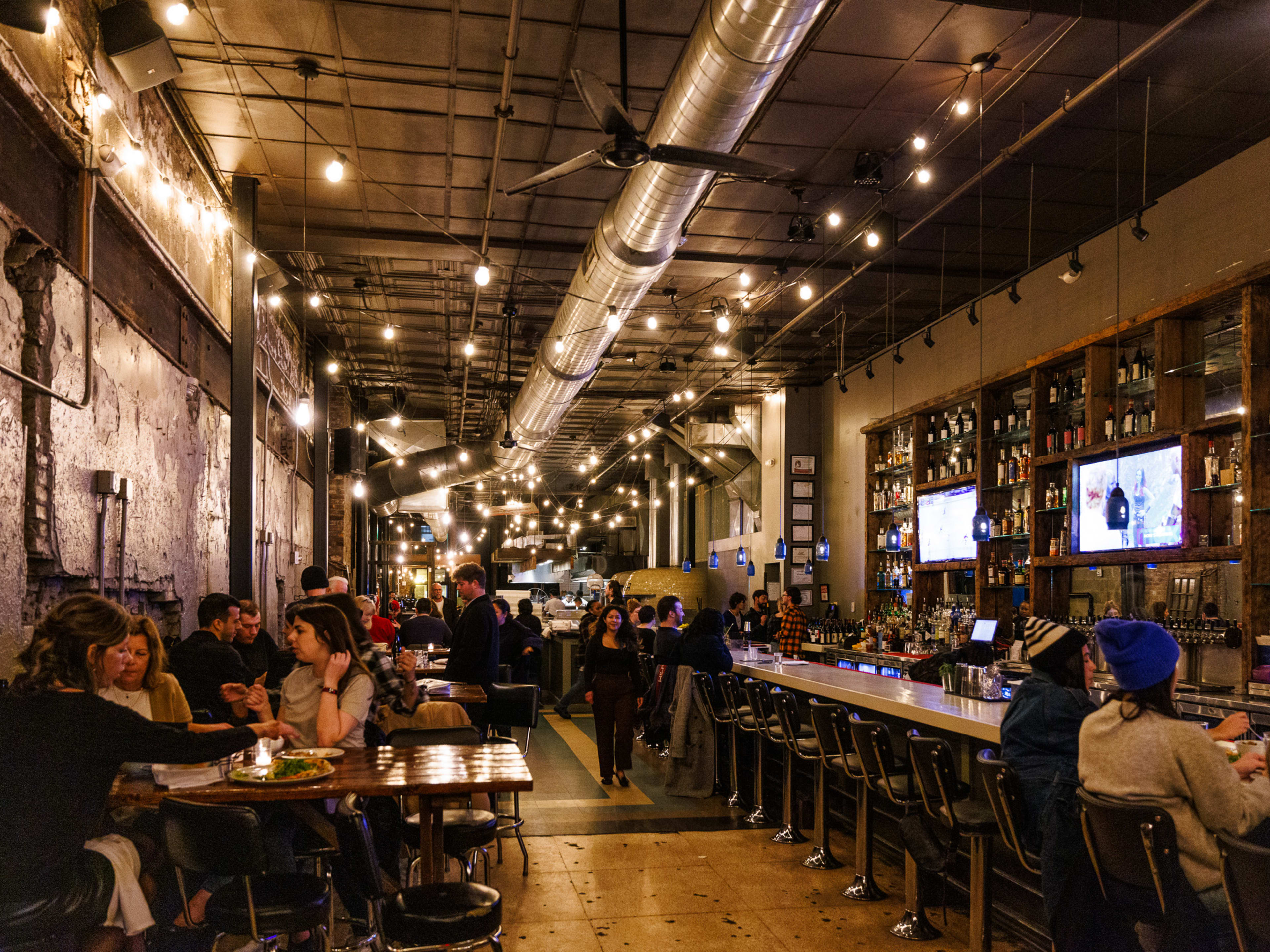 Porta interiors at night with people seated at high top wooden tables on the left, people seated at the bar on the right, and string lights hanging all across the ceiling.