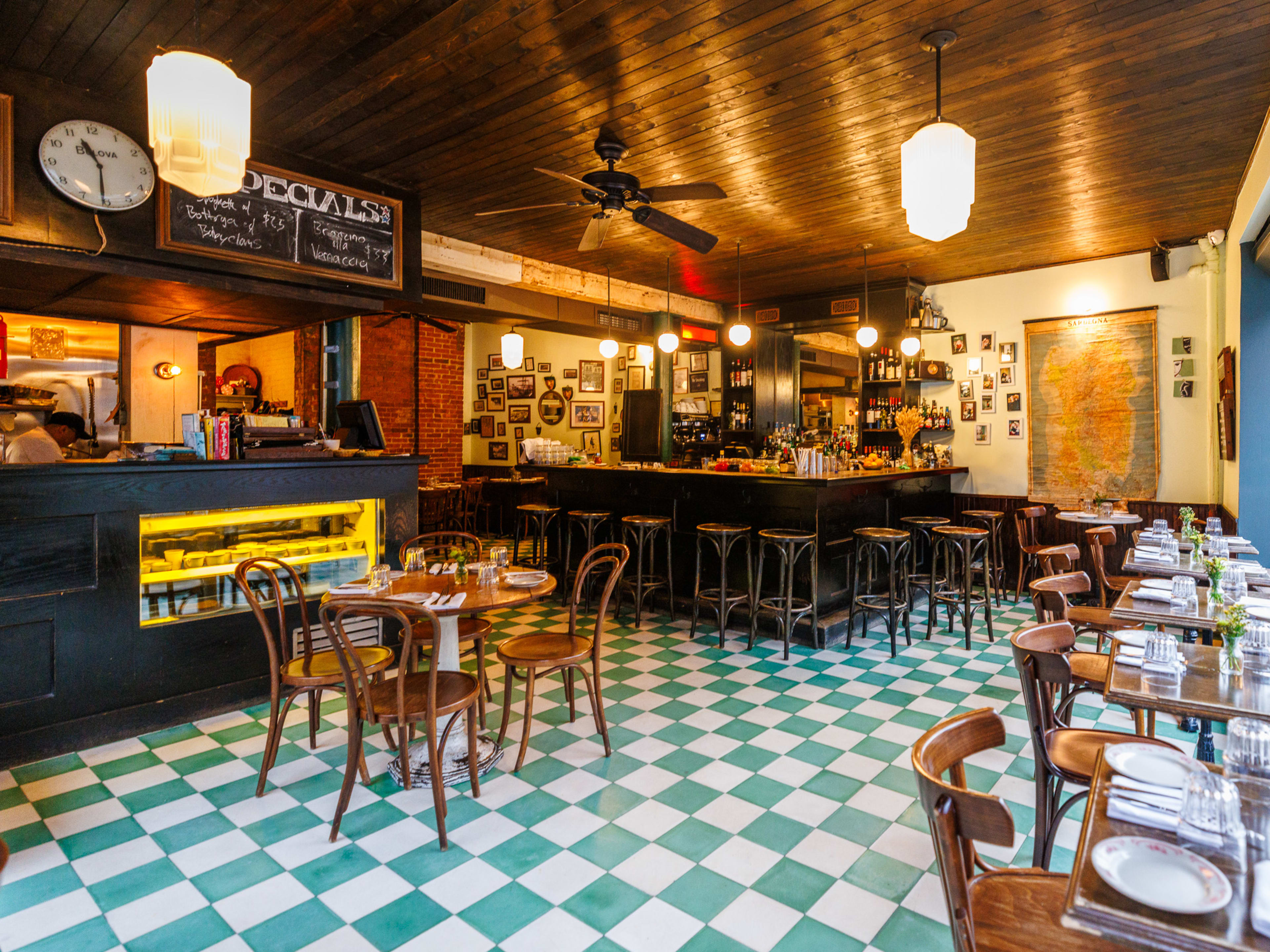 River Deli interiors with green and white checkerboard floors, a wood paneled ceiling, bar stool seating in the back and wooden tables and chairs with place settings and flowers on each one.