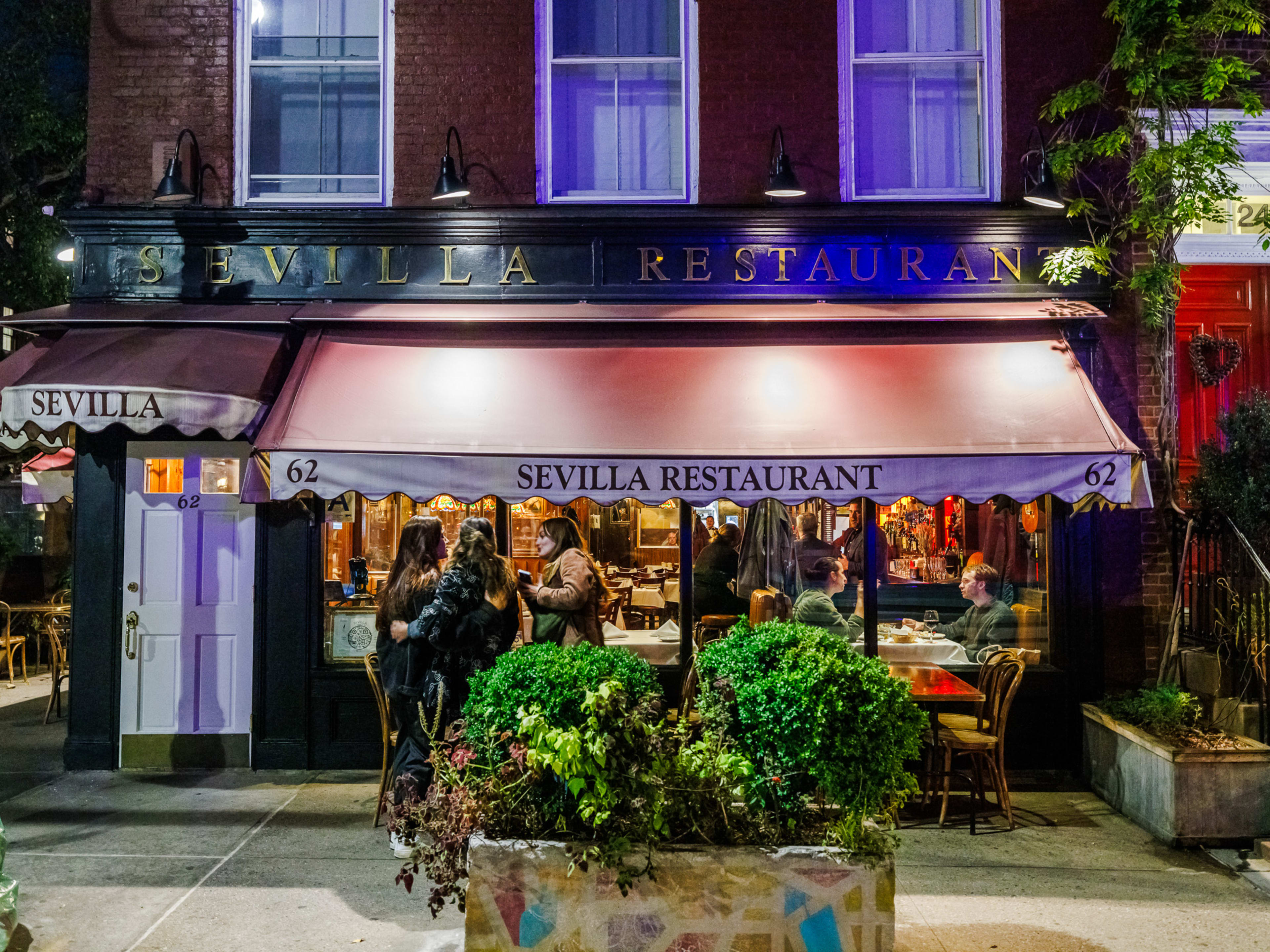 Sevilla Restaurant exterior at night with a large awning, green trees out front on the sidewalk, and people standing by the sidewalk seating area